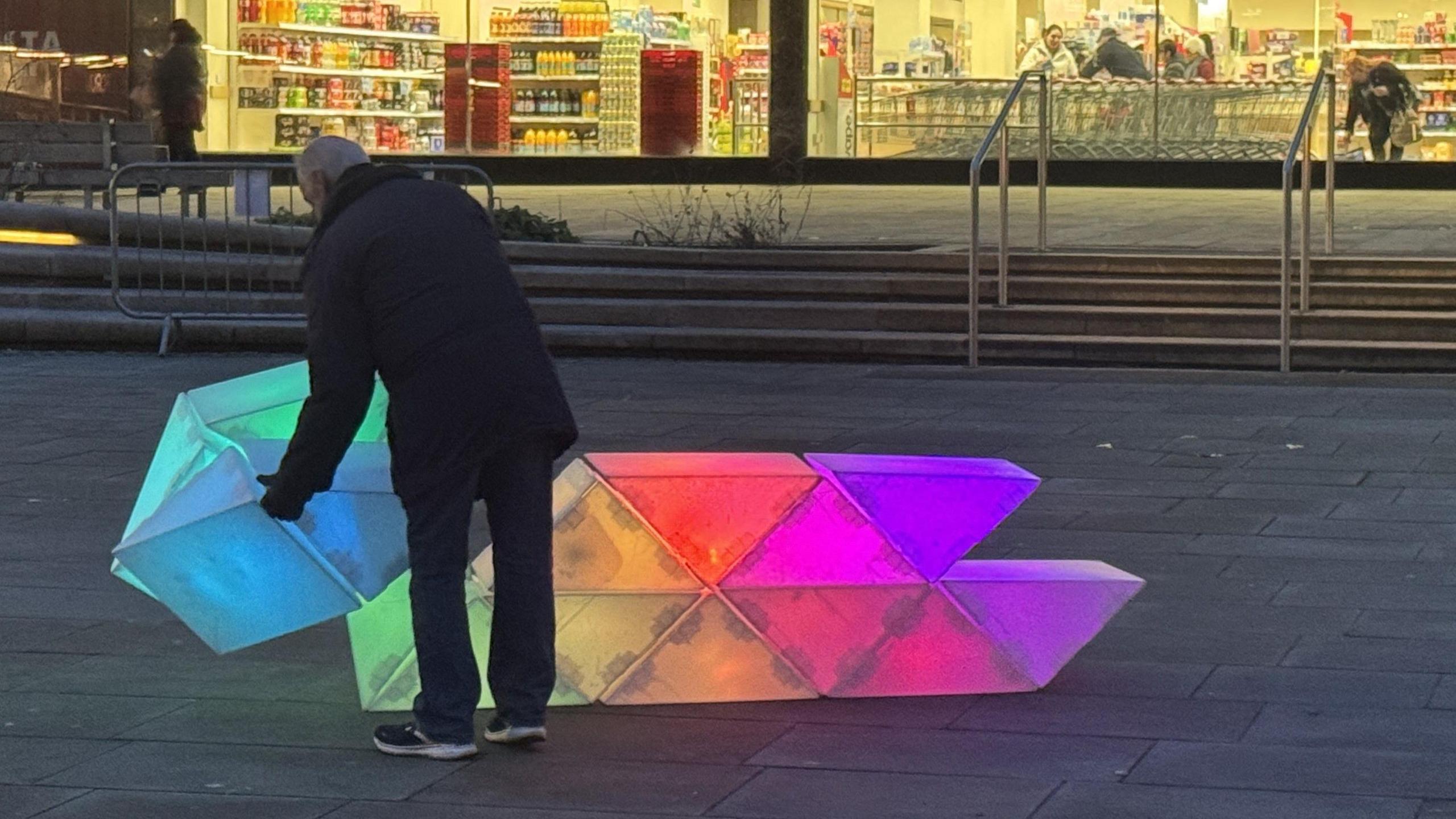 A man stands holding large colourful series of blocks which are illuminated from the inside. They are part of an installation in the centre of Gloucester