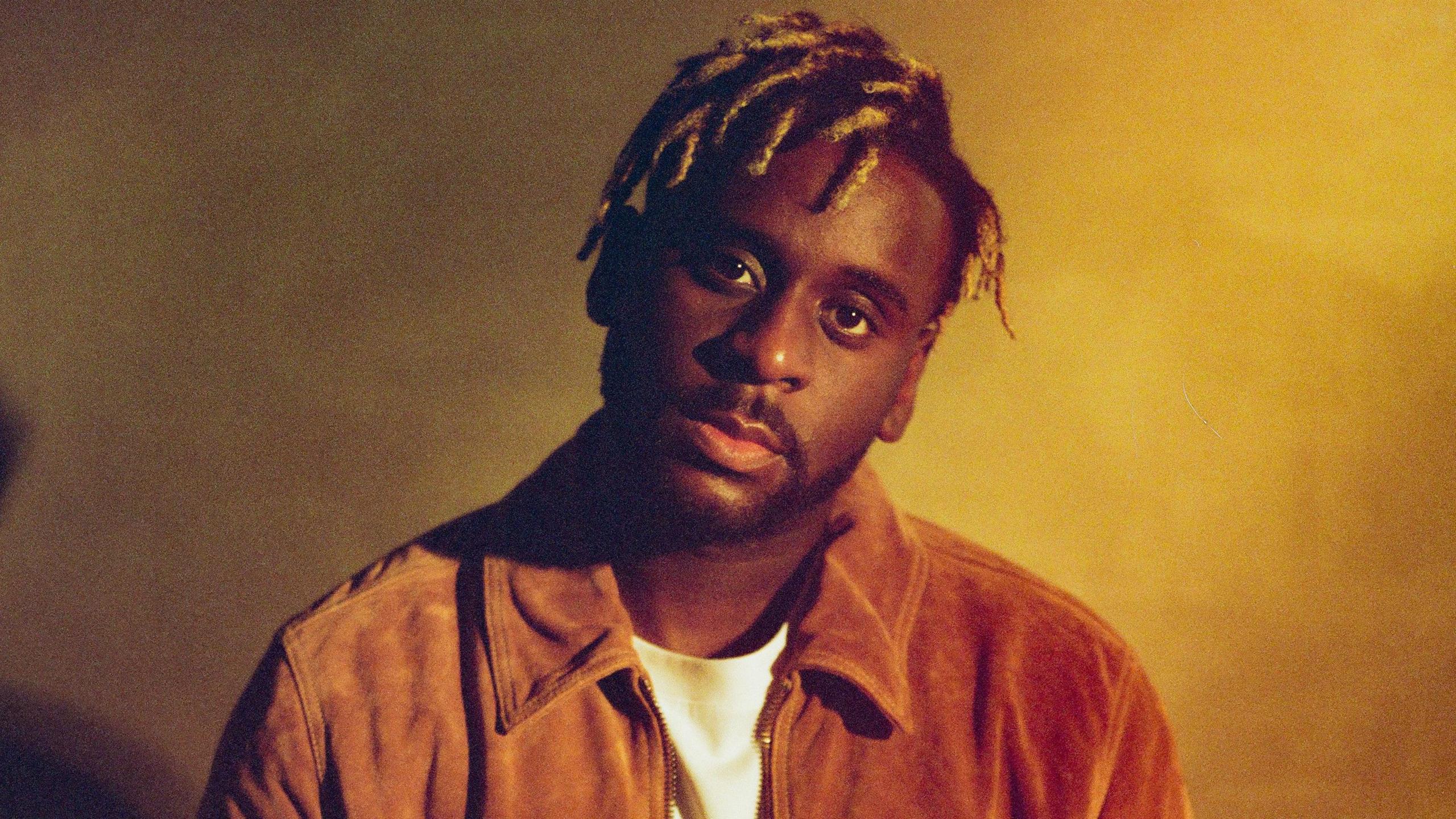 Myles Smith. The singer wears a suede jacket over a white T-shirt and tilts his head to the right. He has a trimmed beard and a serious expression and is pictured in front of a yellow backdrop. 