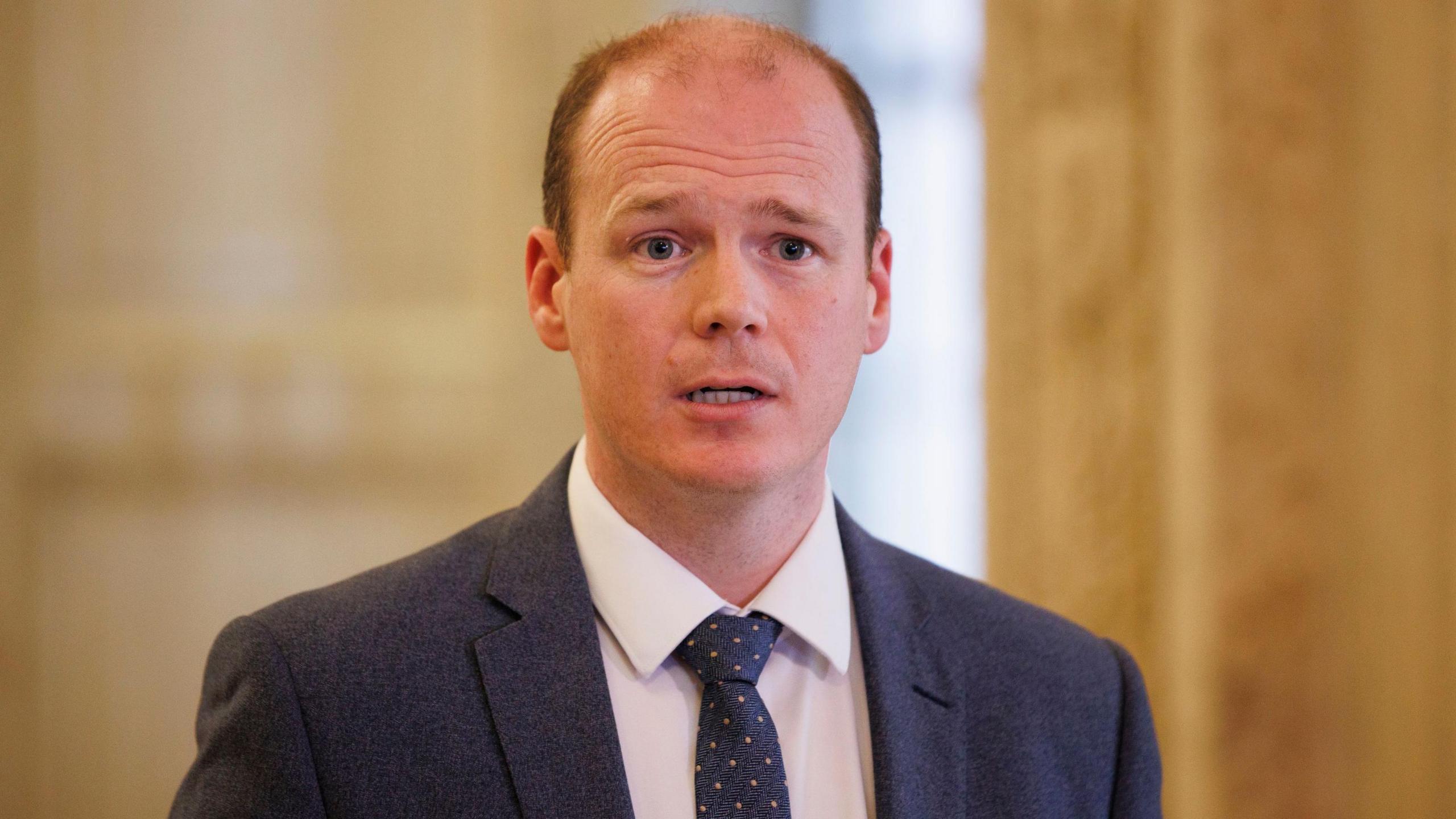 A man wears a grey suit, white shirt and grey tie, with orange polka-dots. Behind him is a yellow wall
