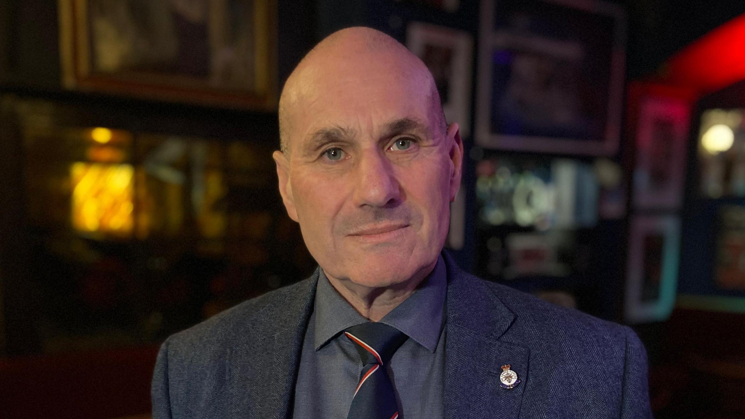 Royal Navy veteran Lt Cdr Duncan Lustig-Prean looks at the camera. He has a shaved head and is wearing a dark grey suit jacket, grey shirt, and black tie with red and white stripes. On his right lapel is a Royal Navy badge. 