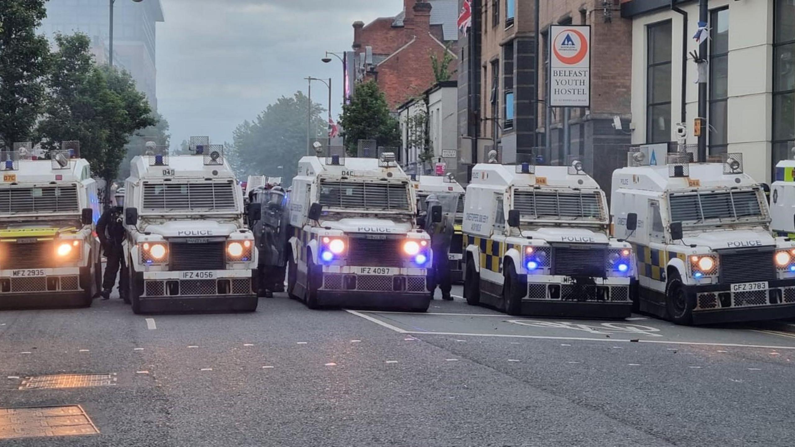 A row of police land rovers lined up across the width of a road with police in riot gear beside them