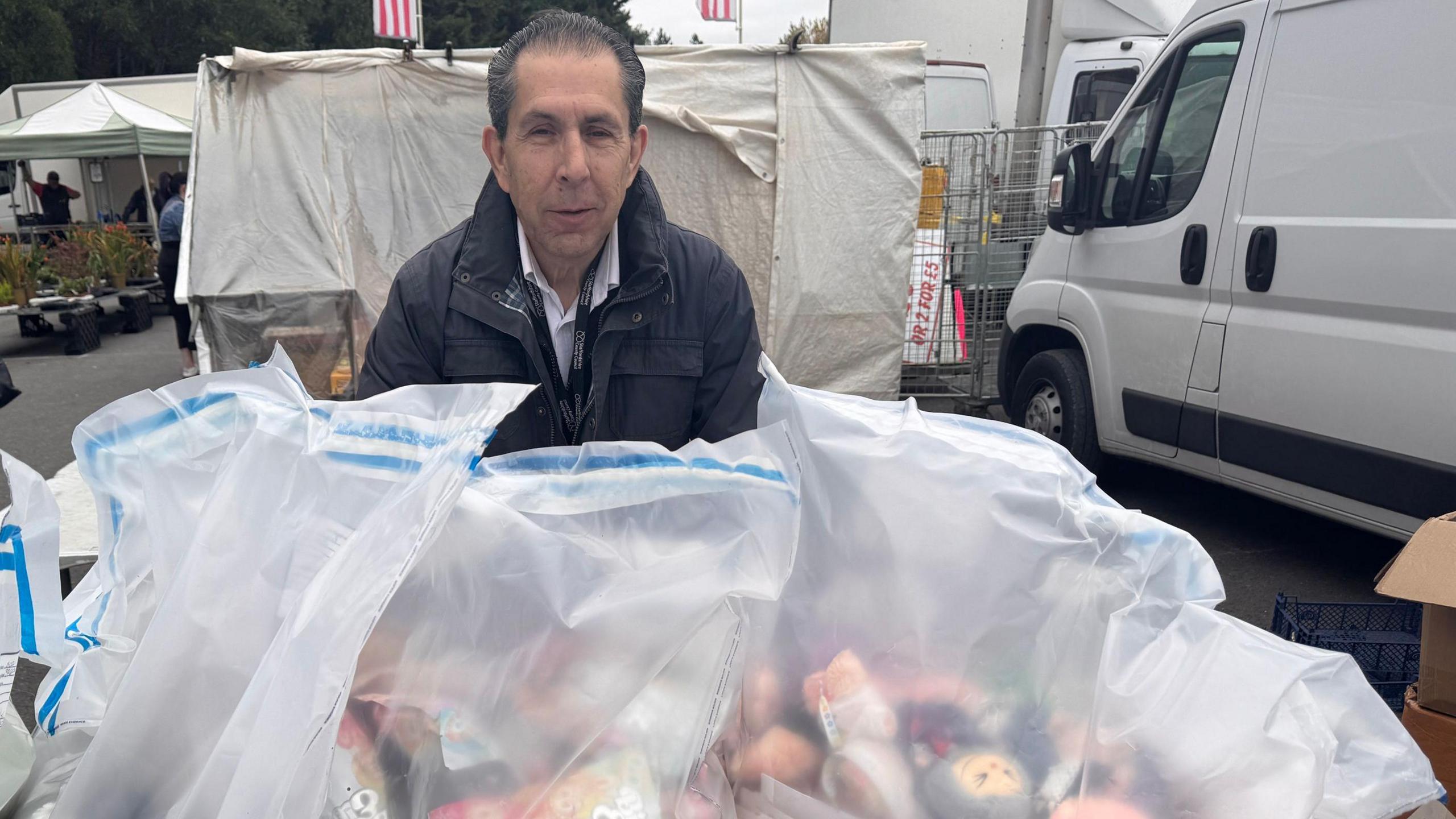 A man stands behind a number of large clear plastic bags containing colourful counterfeit dolls. A van is parked on the right and people can be seen milling around a market in the background.