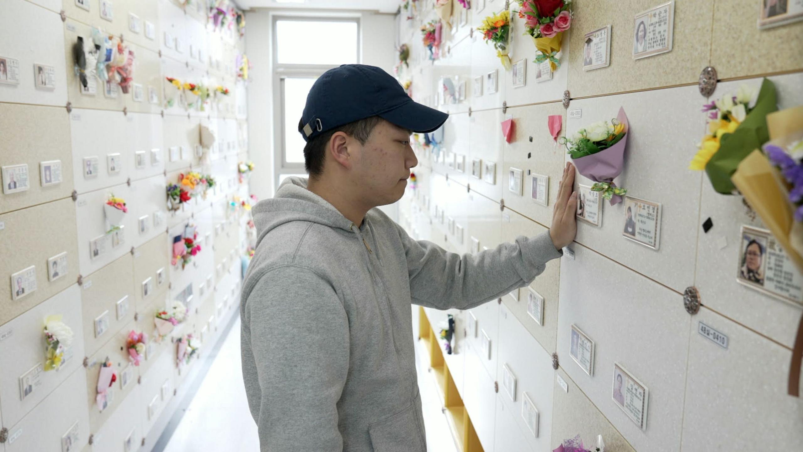 A man in a grey hooded sweater and a baseball cap stands in a memorial, surrounded by garlands of flowers, and holds his hand to a small picture on the wall