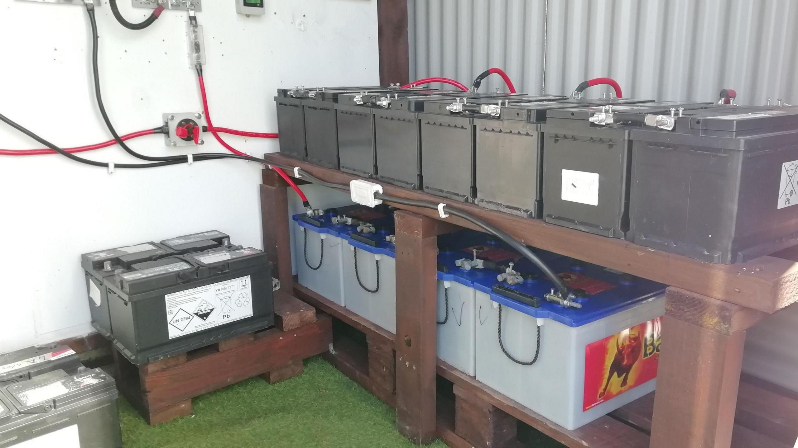 A battery bank at the Wilkinson's home in County Kilkenny, Several large batteries are stacked on wooden shelving.  They are connected to power sources with cables.