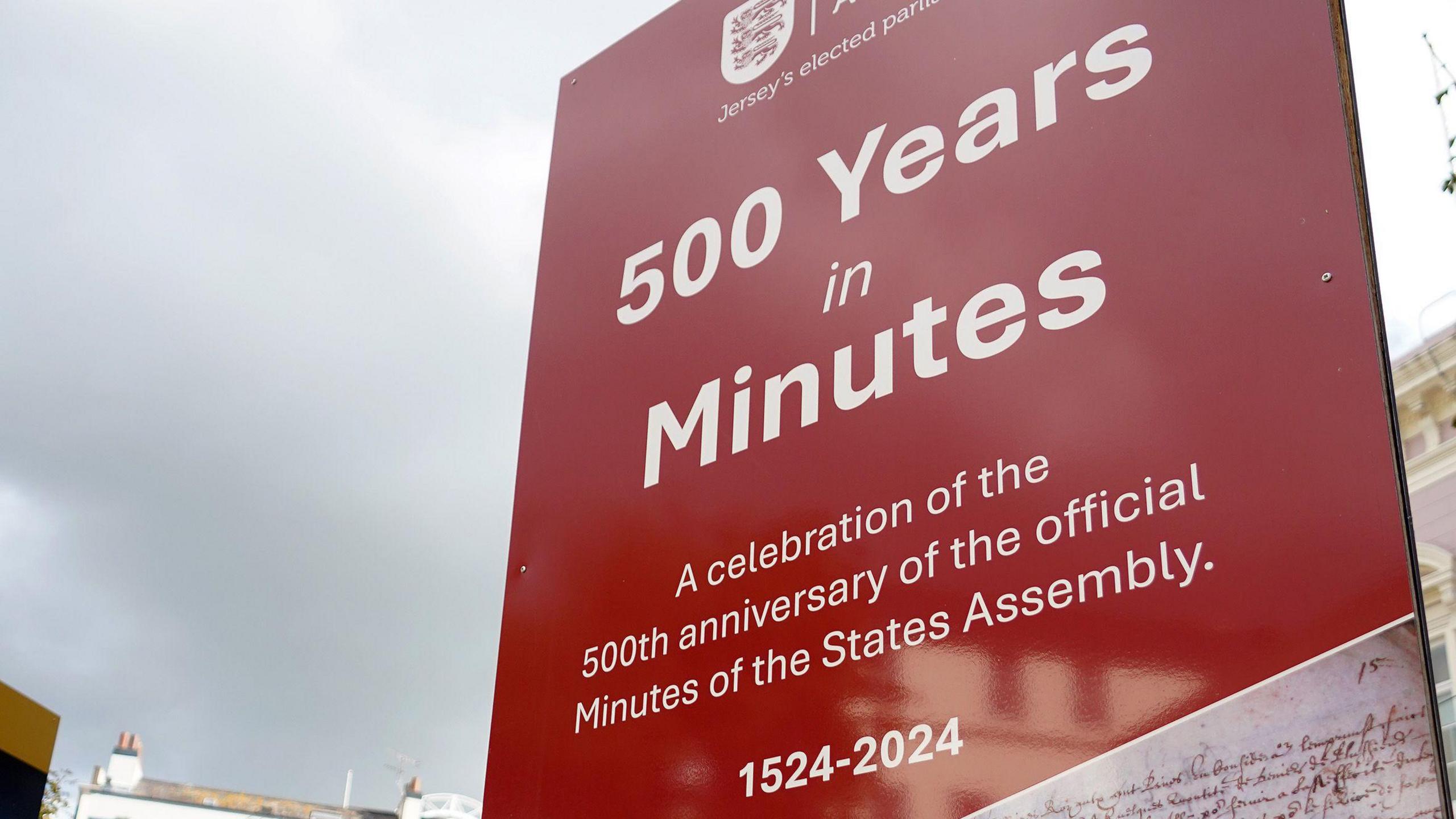 One of the exhibition boards. It has a red background with white writing it says. 500 years in minutes - a celebration od the 500th anniversary of the official Minutes of the States Assembly. A grey sky is visible in the background.