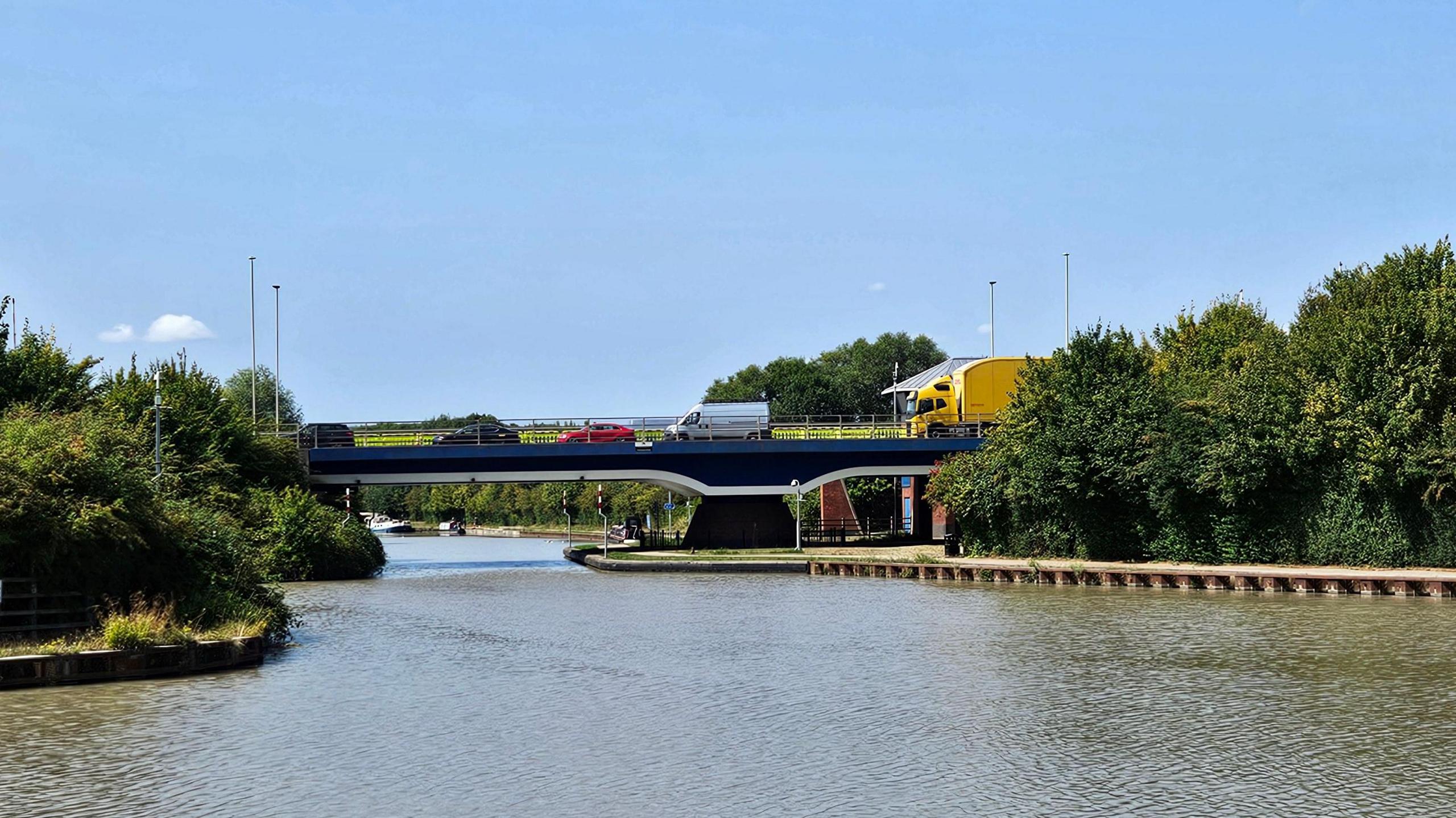 A bridge carries traffic, including a bright yellow lorry, a van, and three cars, over a canal on a sunny summer's day. There are trees on either side of the canal and canal boats can be seen in the distance on the other side of the bridge. There are hardly any clouds in the sky.