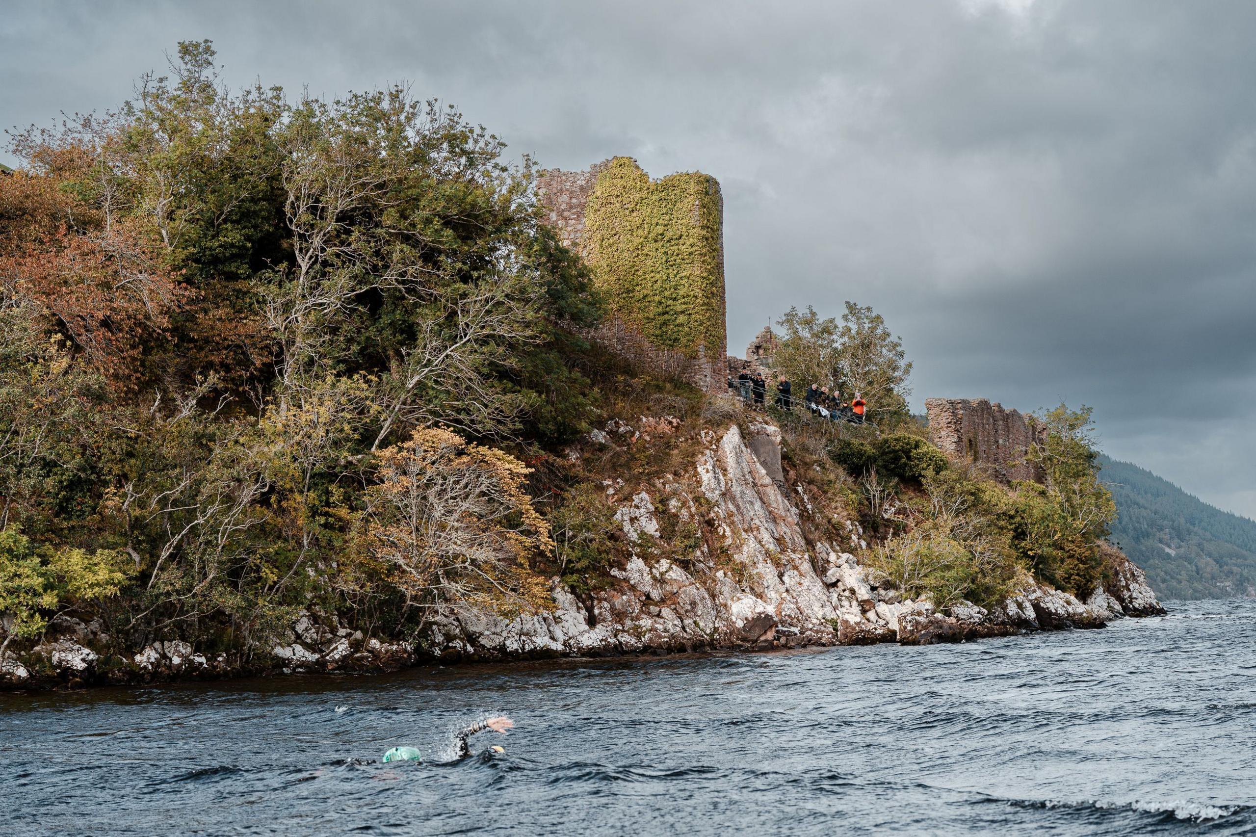 Iain swims through choppy waters of Loch Ness under the ruins of Urquhart Castle. The castle is on a tree covered, rocky promontory. 