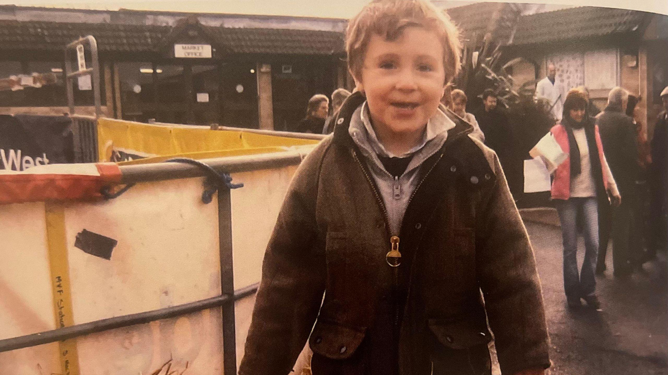 Harry Churchill as a young boy at a farm event. He is wearing a dark zip up top with around three layers beneath of grey, navy and white in colour. He is smiling at the camera and is around aged 6.