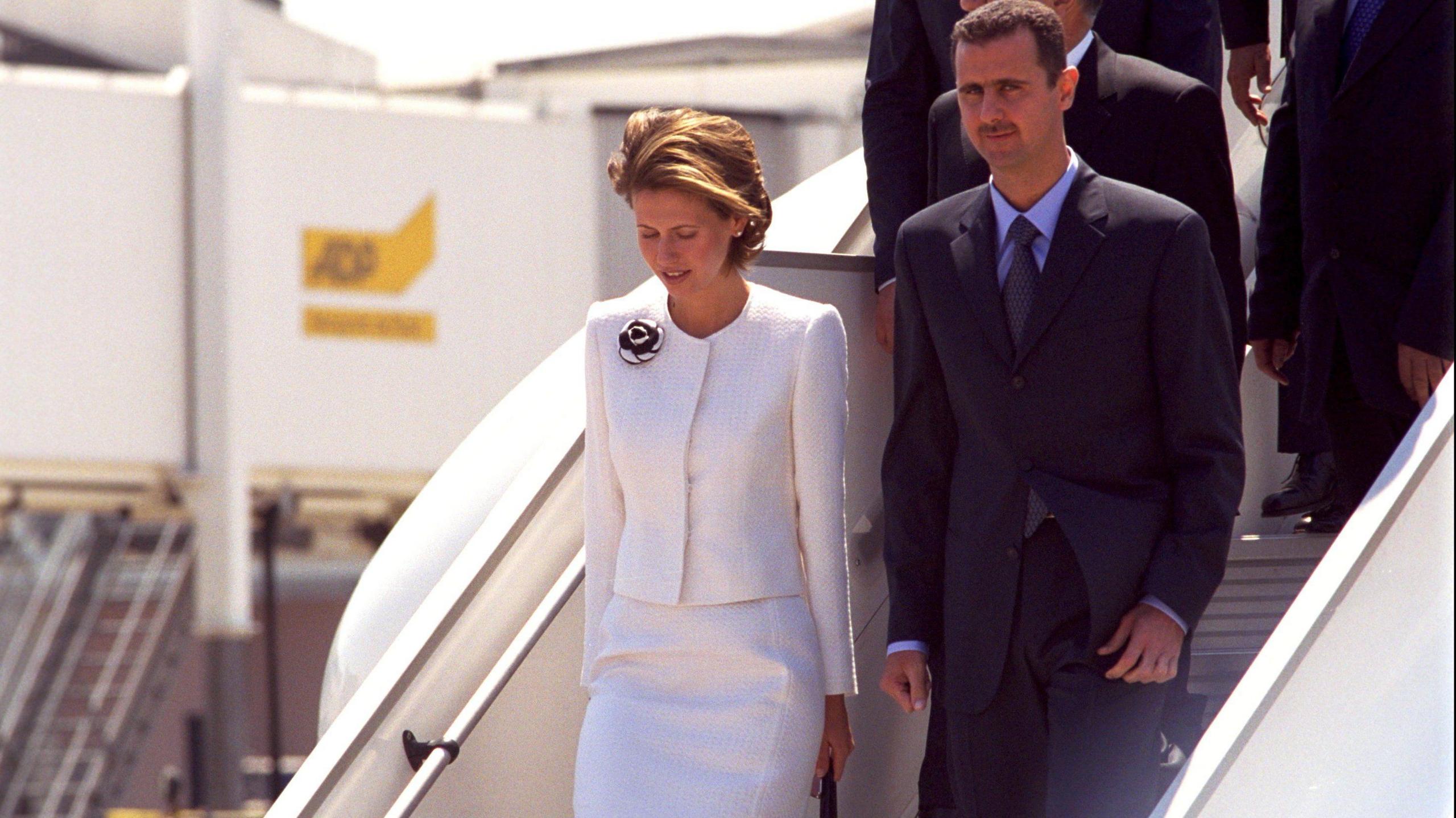 Asma al-Assad in white and Bashar al-Assad in black descend a plane staircase.