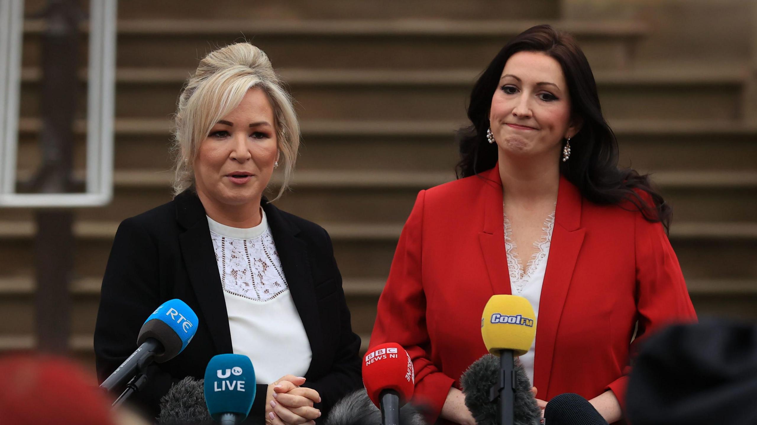 Michelle O'Neill and Emma Little-Pengelly are standing side by side in front of media microphones. O'Neill has shoulder length blonde hair. She is wearing a white blouse and a black jacket. Little-Pengelly has long dark hair. She is wearing a white blouse and a red jacket.