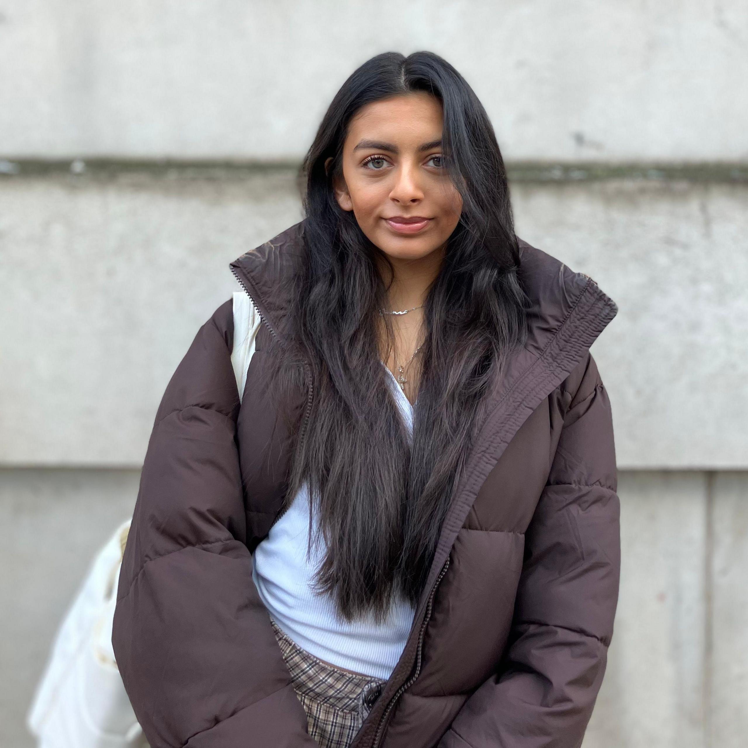 A woman wearing a dark brown padded coat with a white shoulder bag visible on the left side, standing against a light grey concrete wall. Long dark hair falls over the coat, and a thin silver necklace is visible at the neckline.