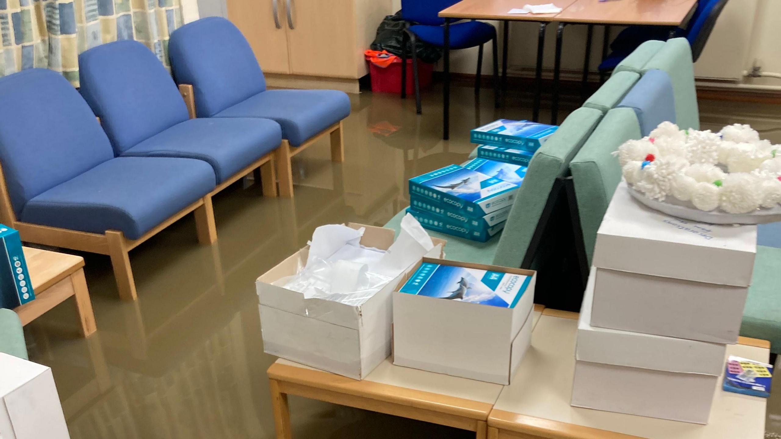 Flood water covers the floor of a school room. A row of three blue chairs stand in the water. Books are stacked on another set of chairs and a table.