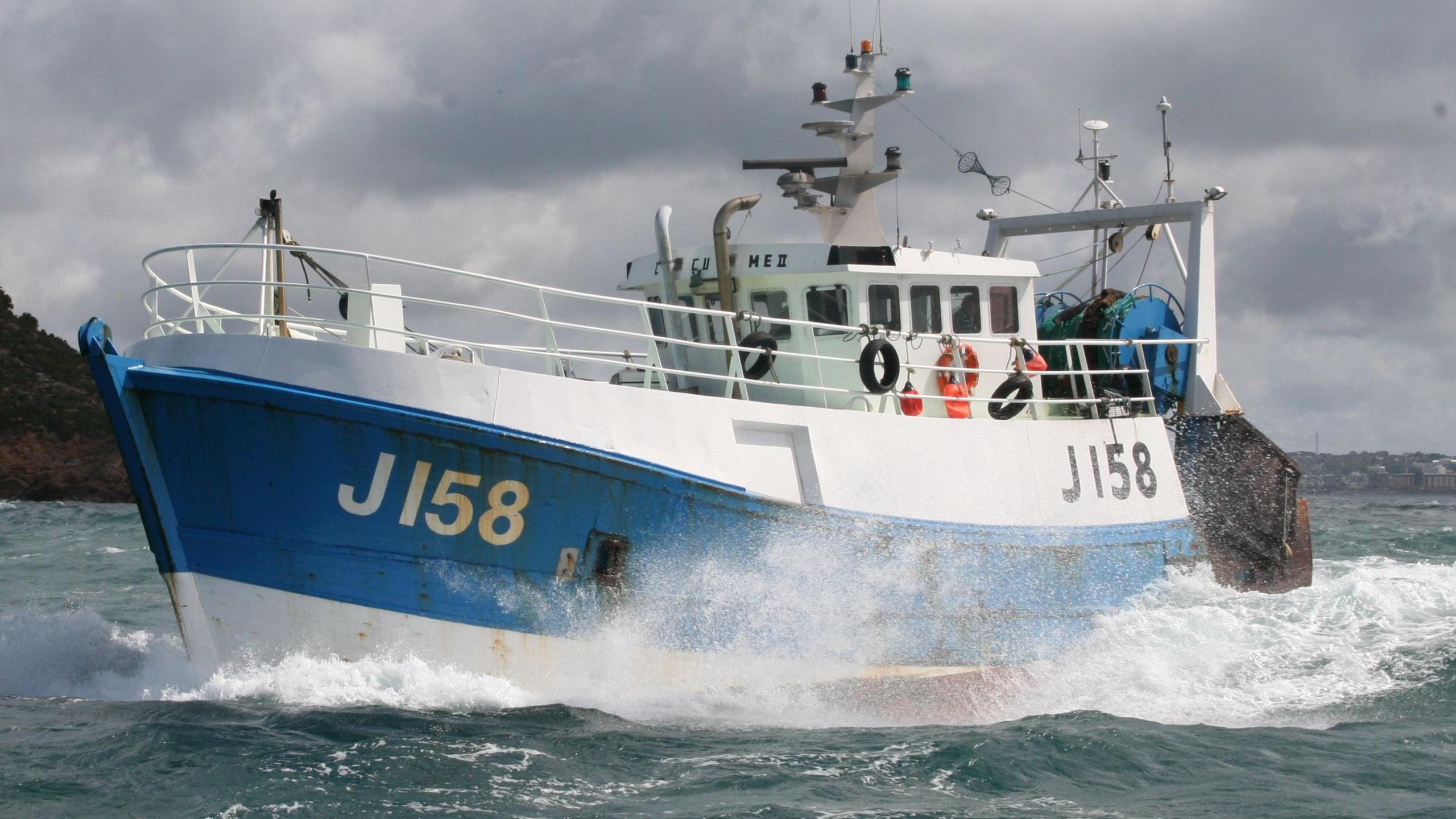 A white and blue fishing boat with J158 on the left side, in the ocean sailing. Land can been seen afar in the background.