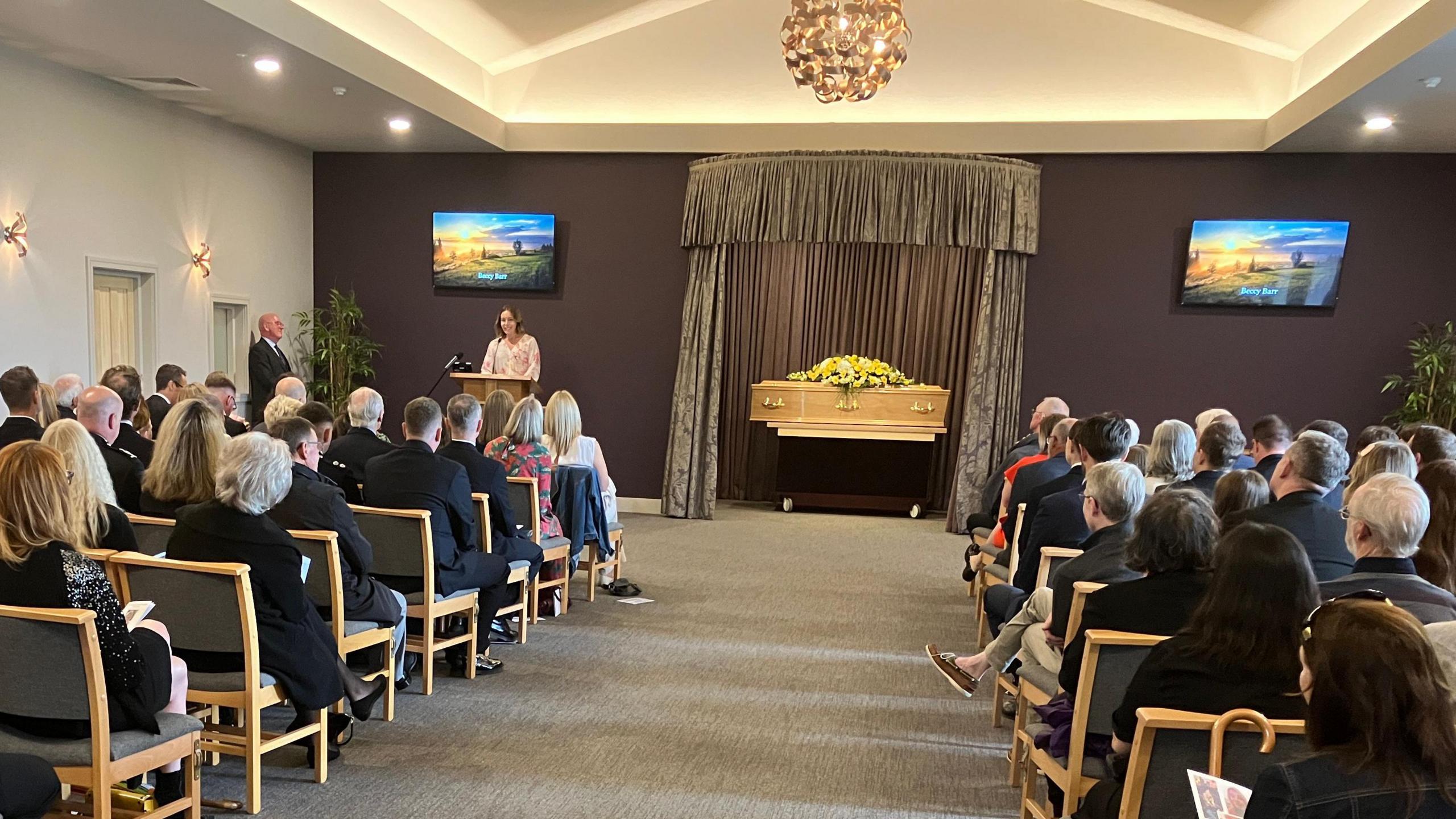 Lorna Mendelsohn gives a eulogy at the funeral of Beccy Barr. She stands at the lectern to the left of the coffin as the camera looks down the aisle between two rows of seated mourners