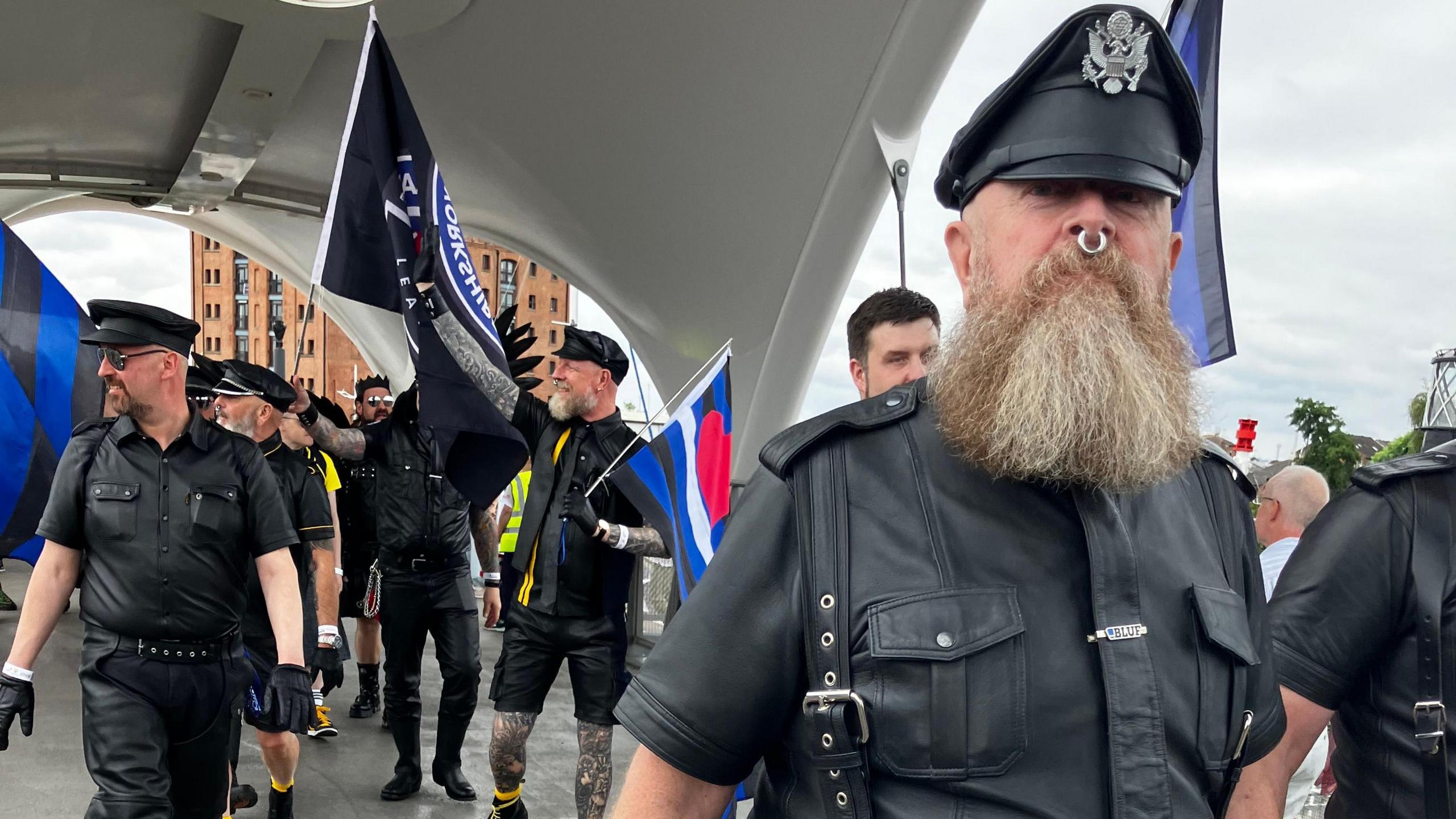 A group of men looking like the Village People, with black leather, black police-type hats, lots of tattoos and studded belts. A man in the foreground has a long grey beard and a large ring through his nose.