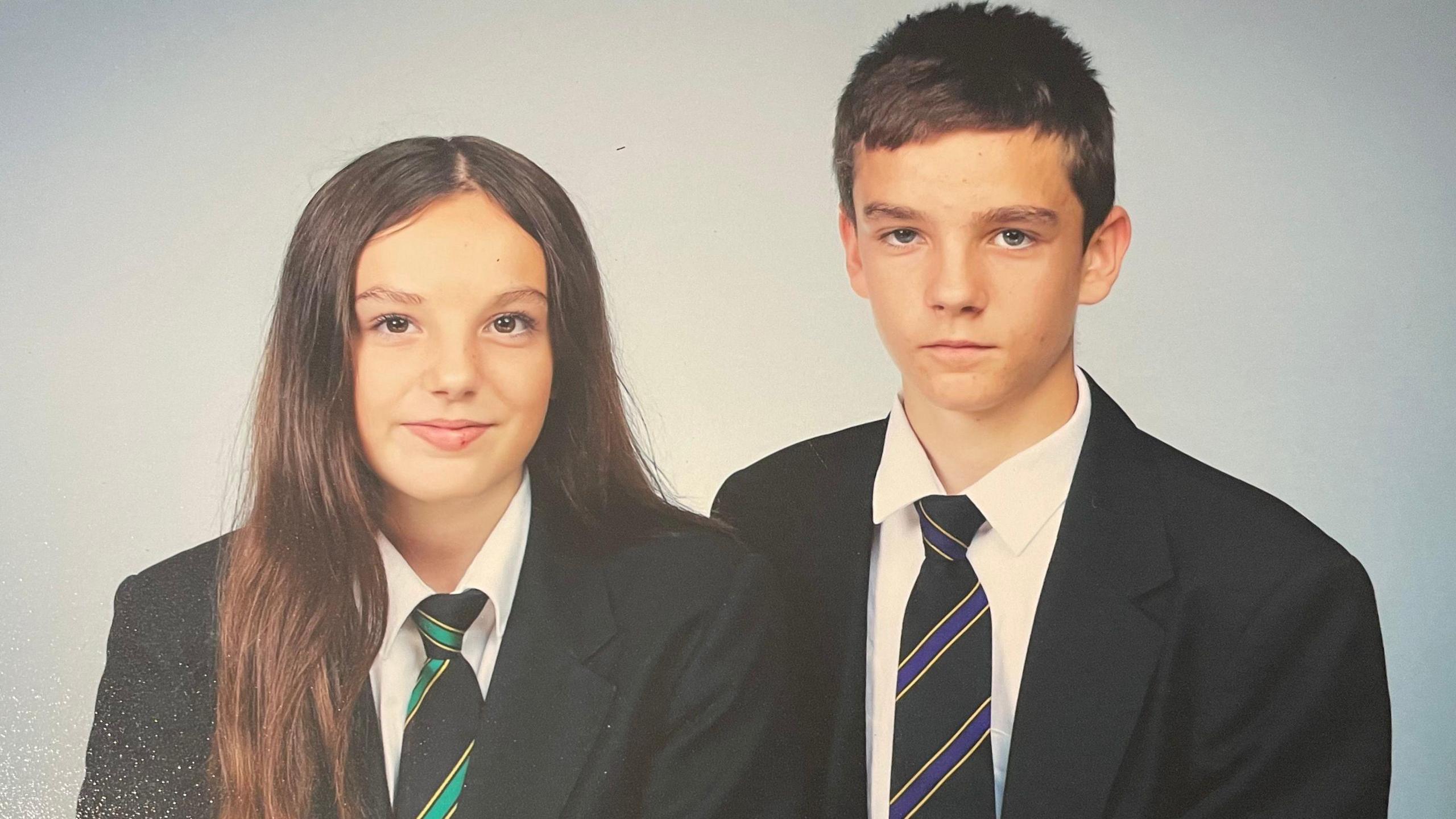 Freddie Coleman and his sister. Both are wearing school uniform, consisting of blazers, white shirts and black ties. Freddie has short, dark brown hair and he is looking at the camera expressionless. His sister has long brown hair and is smiling. The picture has been professionally taken on a light blue background.