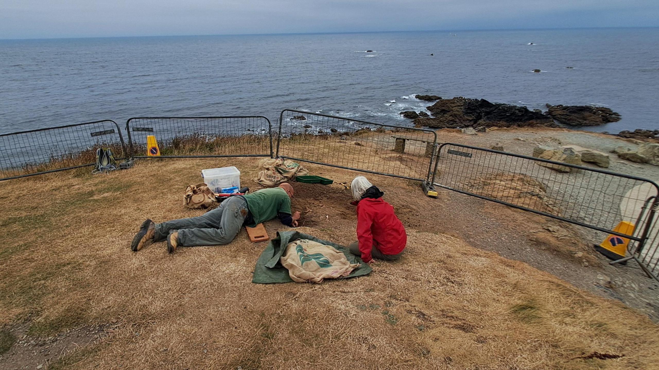 A man is lying down examining the site and a woman is sitting next to him. The grassy area is fenced off and leads to a headland with rocks and the sea beyond.