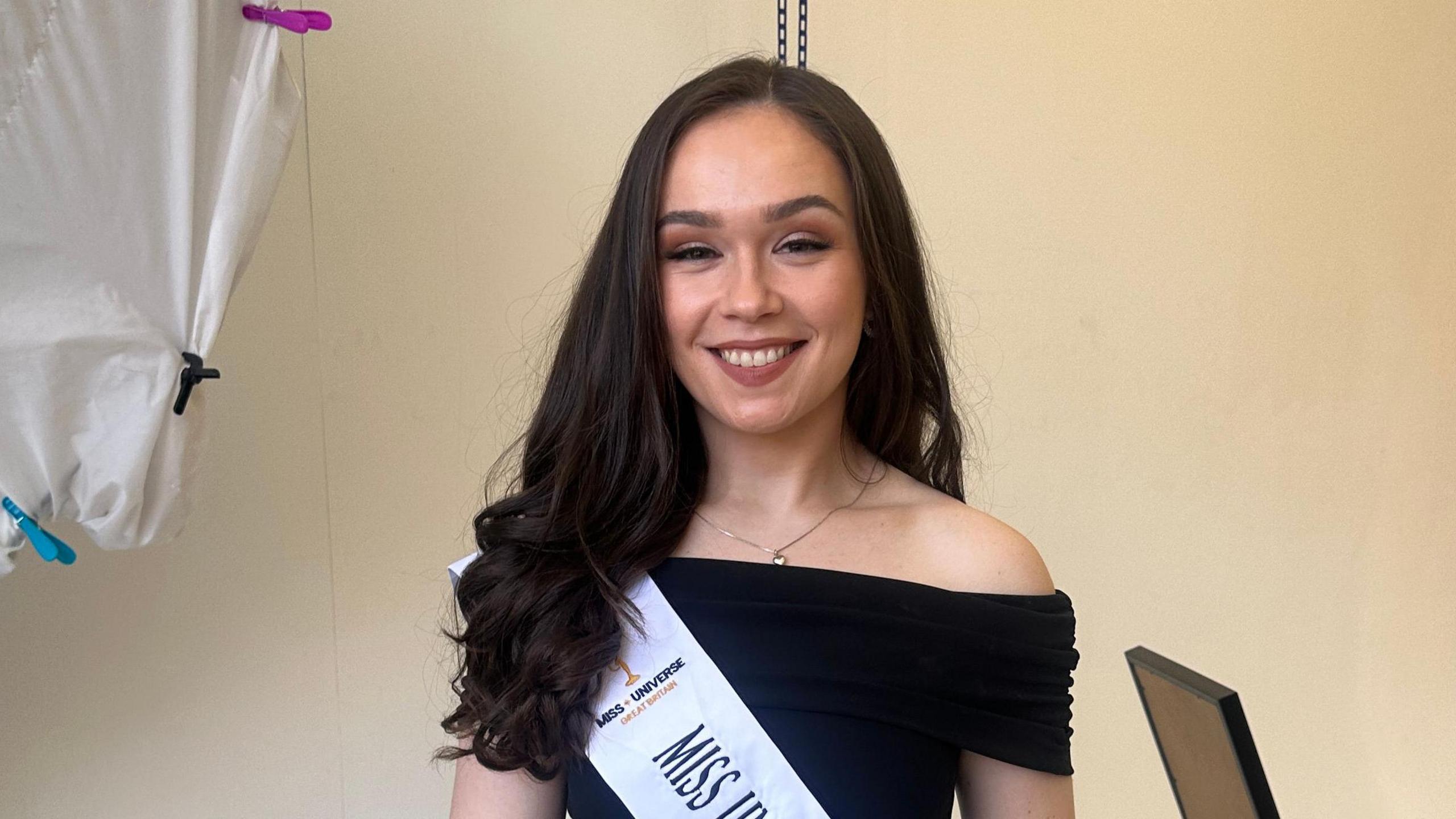 A woman wearing a black dress smiling at the camera. She has red lip stick on and a sash across her chest with Miss Universe Great Britain written on it.