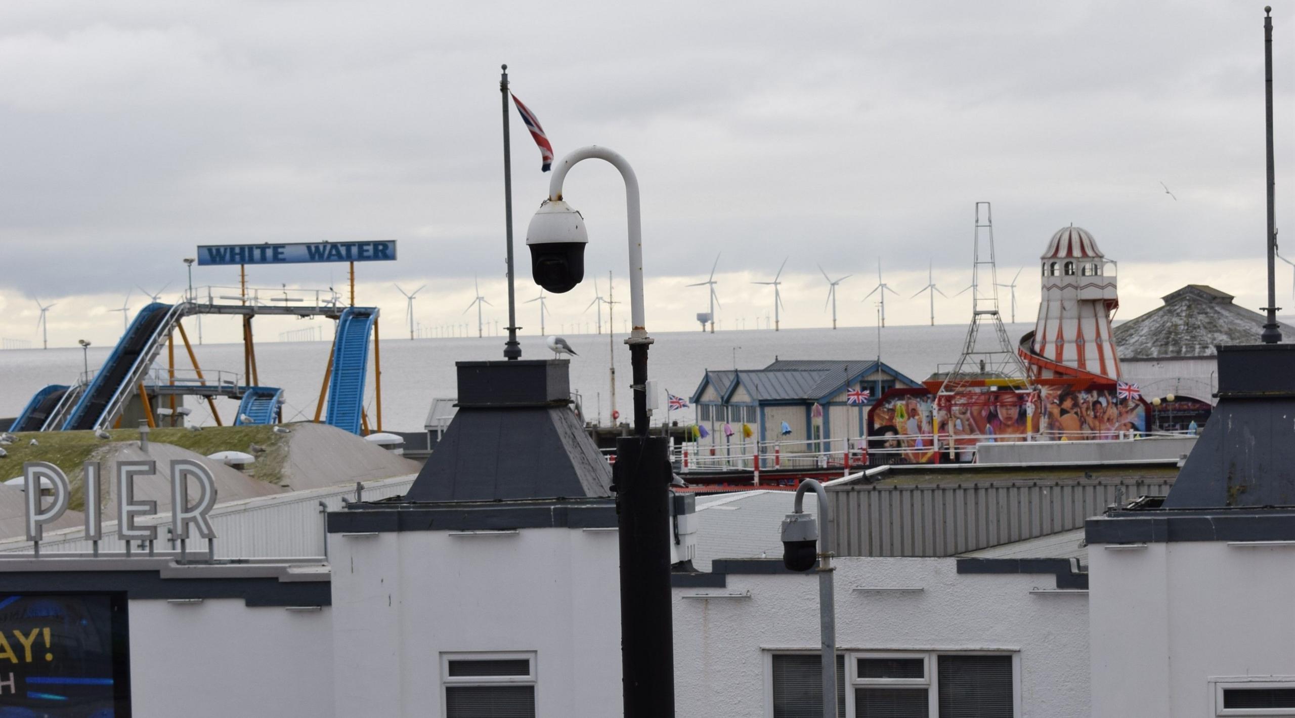 A CCTV camera next to Clacton-on-Sea Pier. The pier is behind it. Off-shore wind turbines can be seen in the distance. 