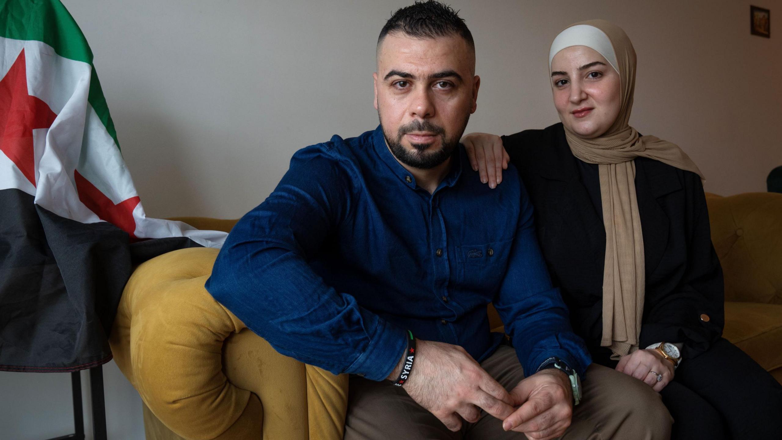 A man in a blue shirt sits next to a woman in a headscarf, her hand on his shoulder.