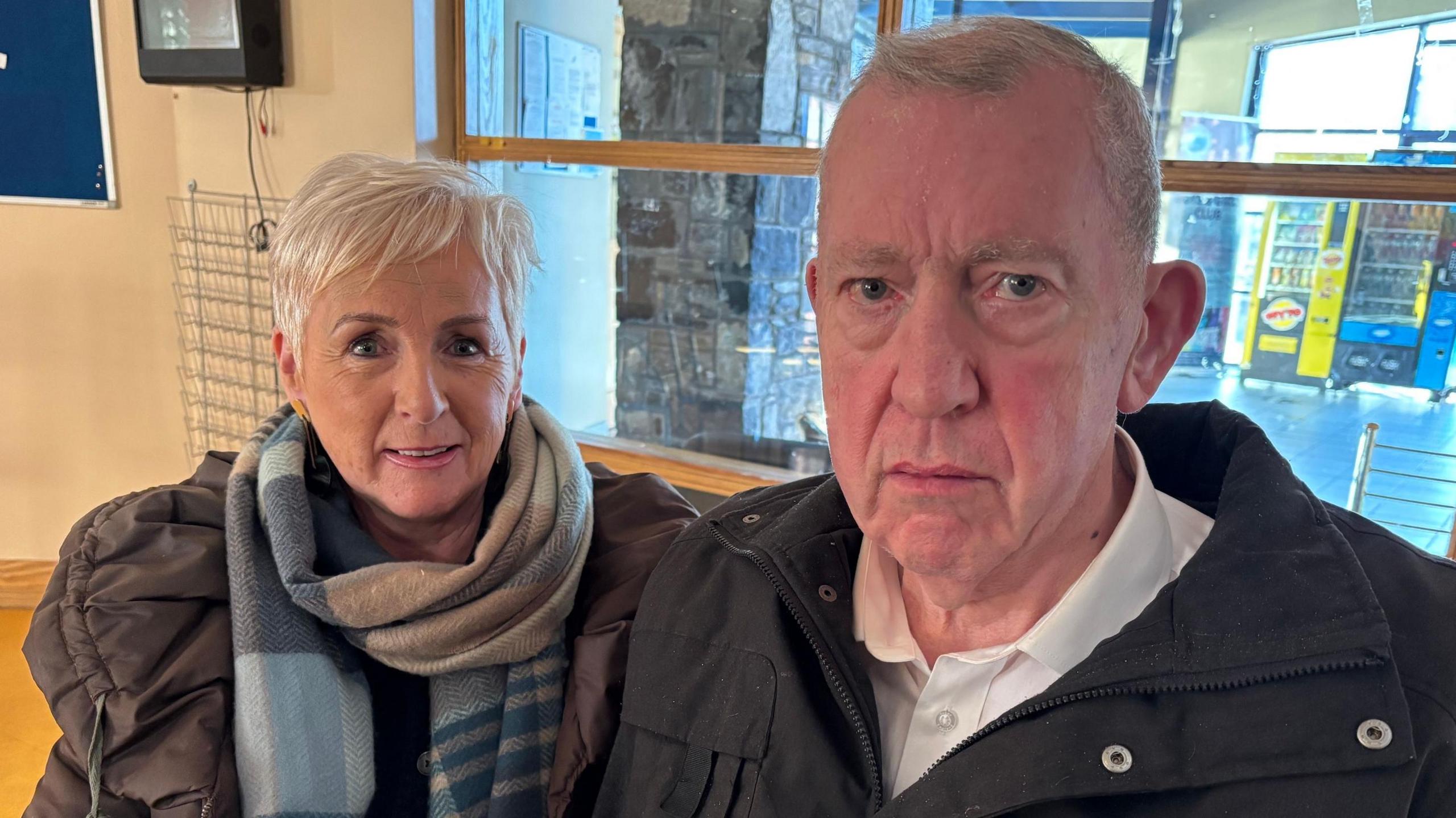 A woman and a man sit together in a leisure centre. They are warmly dressed in coats and a scarf. The woman has short pixie cut grey hair and the man is grey haired with a somber expression.