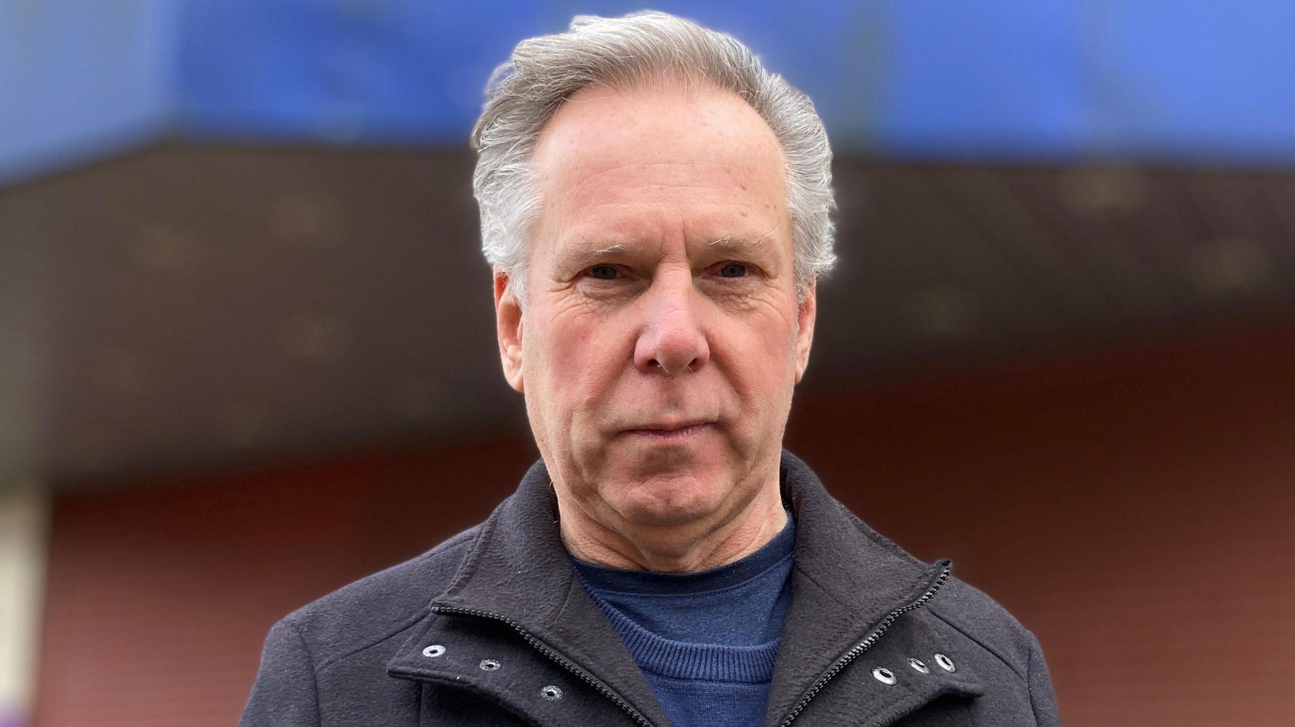 Trevor Wicks, a man looking into the camera. He is stood in front of the Regent Theatre and is wearing a blue jumper and grey coat. 