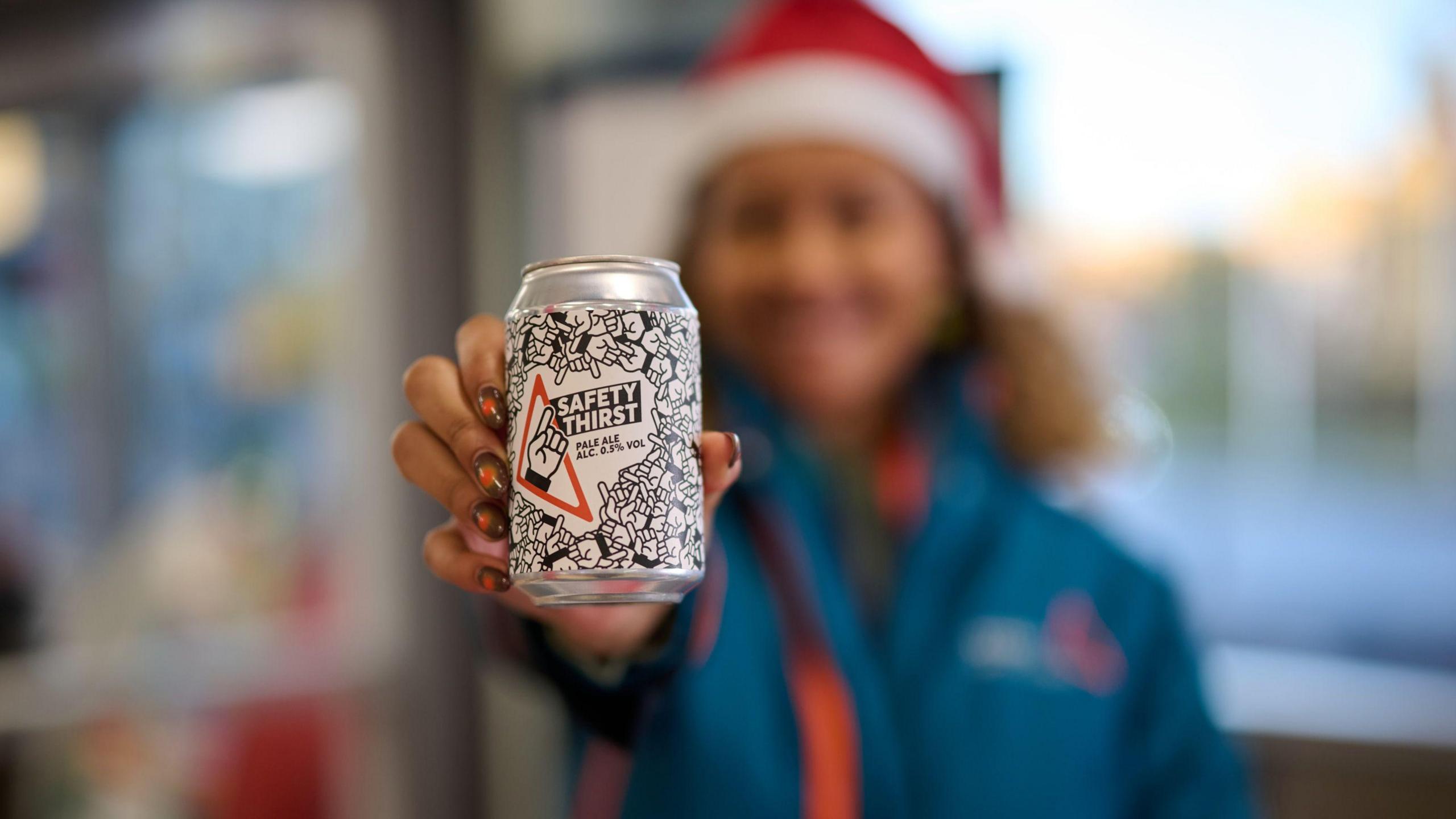 A woman wearing a blue Avanti West Coast jacket and a Christmas hat holds up a black and white Safety Thirst beer can. The woman is blurred and the beer can and her hand are not.