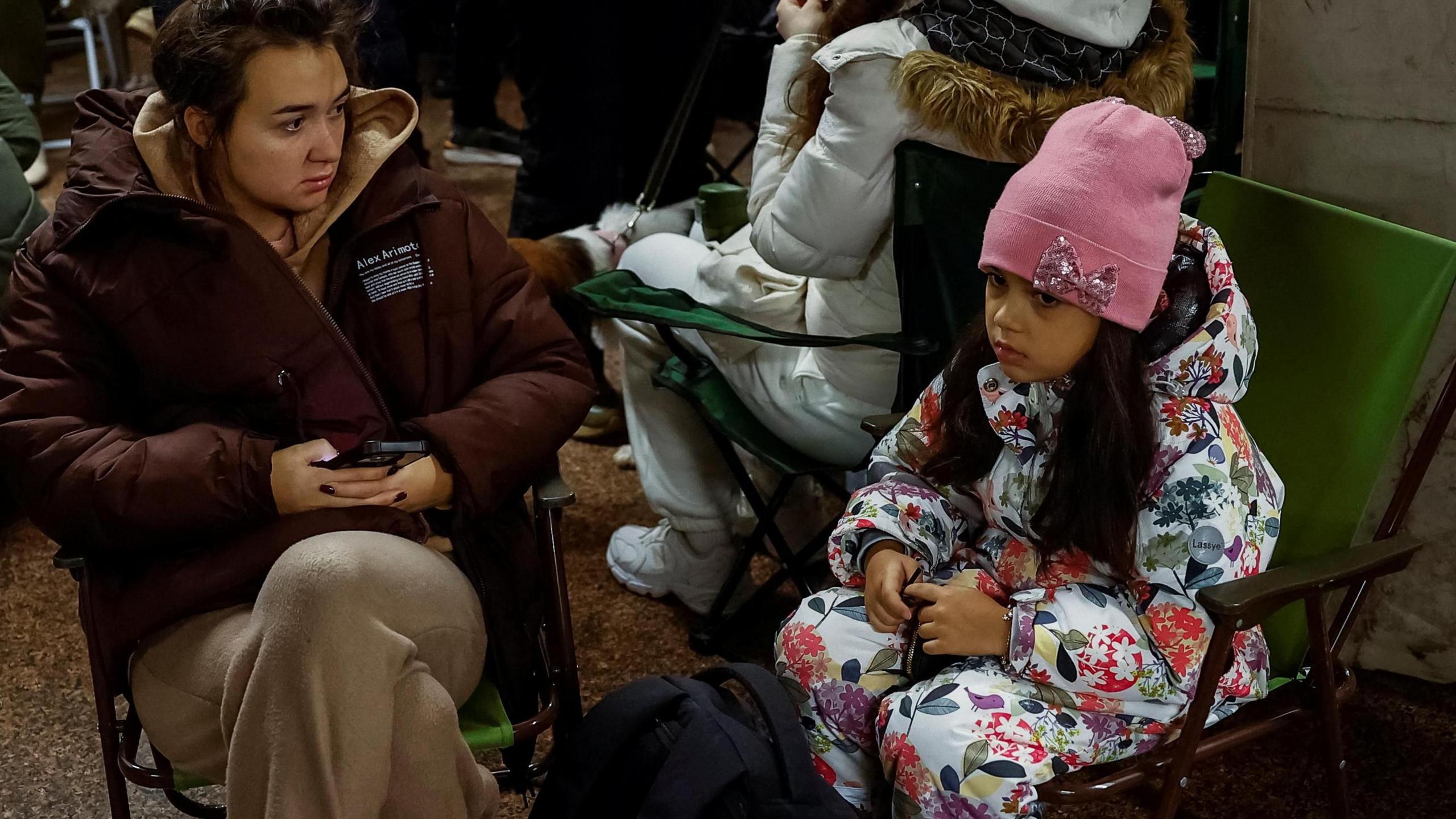 A woman and a young child sit in camping chairs in an air raid shelter in  inside a metro station during a Russian missile and drone attack on Ukraine. The woman has her phone in her hands and both of them look anxious. The young girl is wearing a pink wooly hat and a onesie with a floral pattern on it.