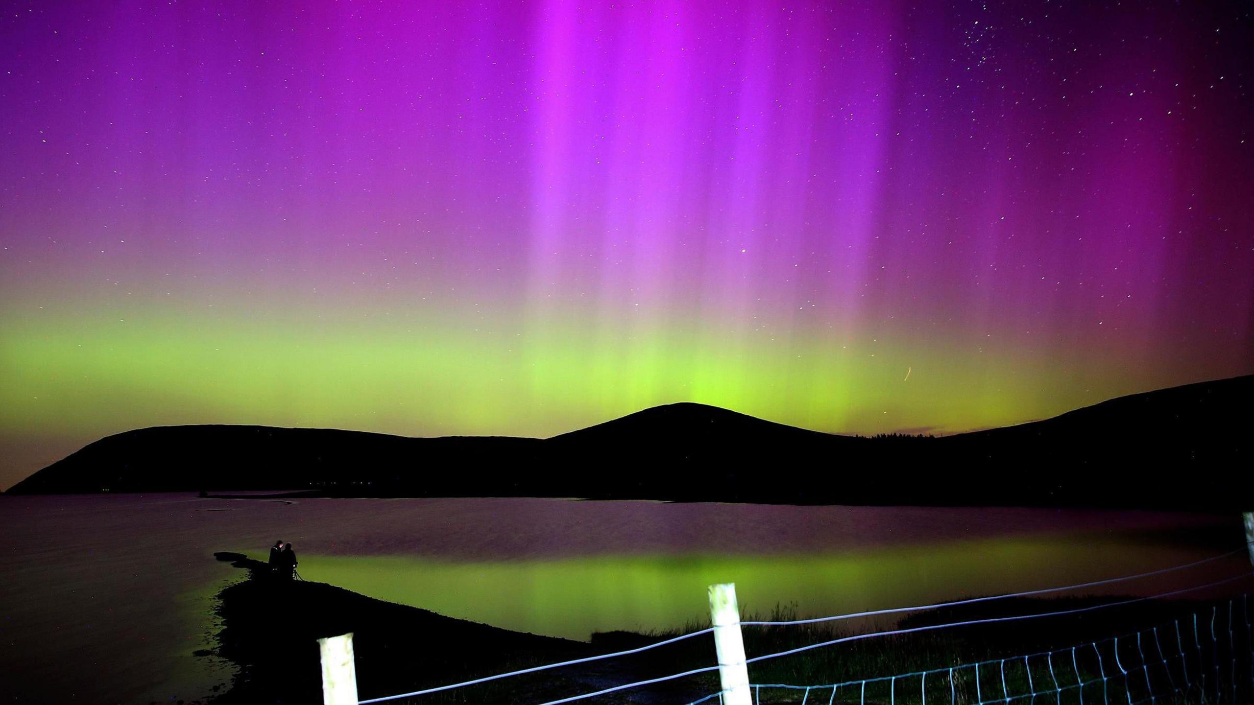 Purple and green northern lights over the silhouette of some hills and reflecting off the sea beneath them 