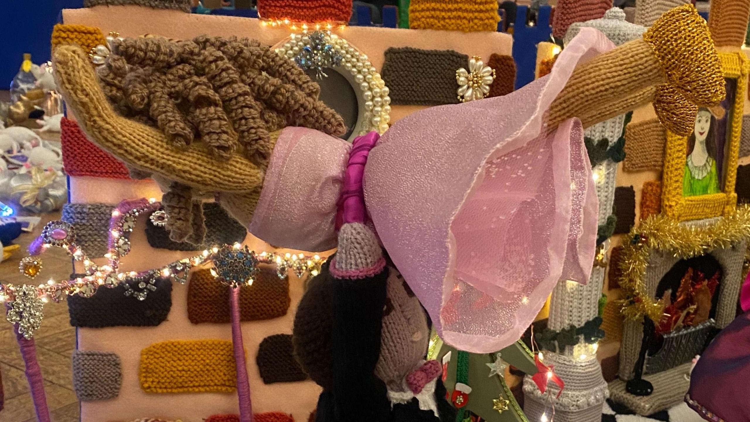 Two knitted dancers completing a lift. The woman is wearing a pink dress and has curly hair. The man is holding her up from her waist. You can see lights behind and a wall. 