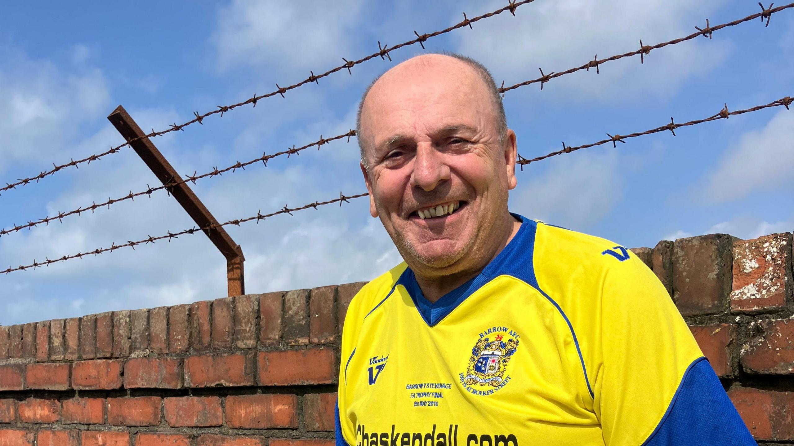 Mr Steele is wearing a yellow Barrow AFC shirt. He has very short hair and is smiling at the camera.