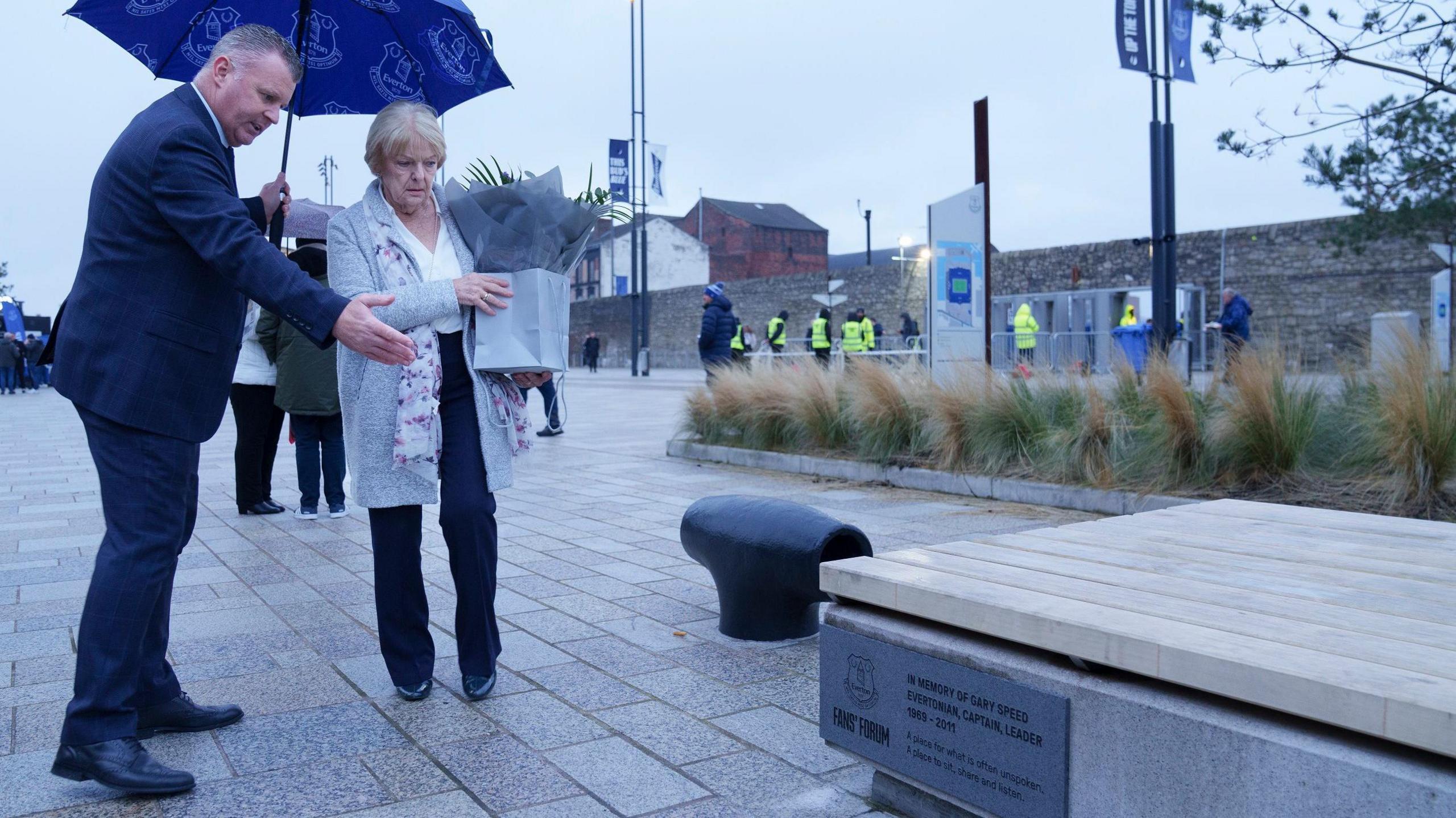 Graham Stuart shows the bench to Carol Speed as she holds a bouquet of flowers. Fans and workers in hi-vis vests mill around in the background.