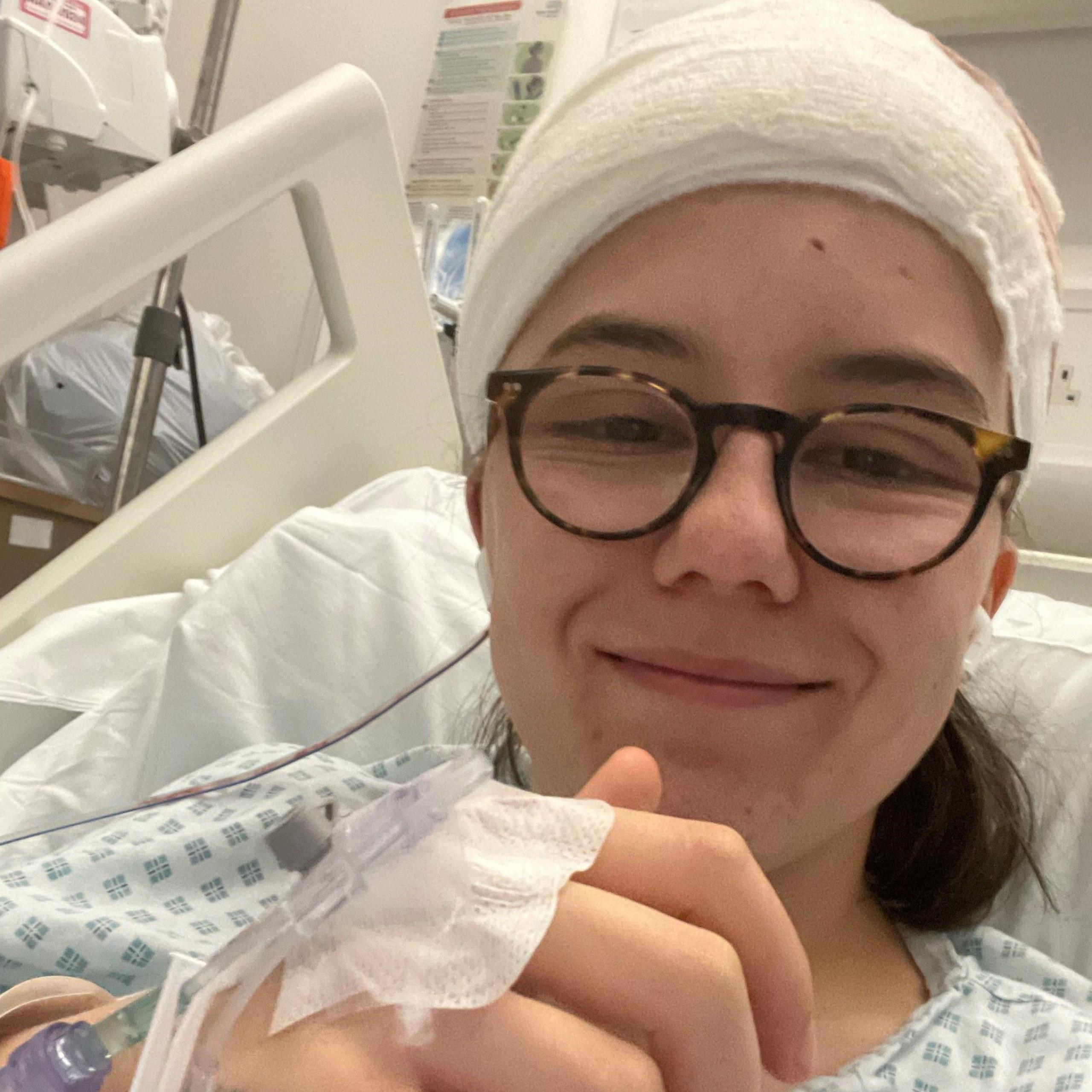 Jesse is seen smiling in a selfie from her hospital bed. She is wearing glasses and her head is bandaged and iv drips are seen in her hand.