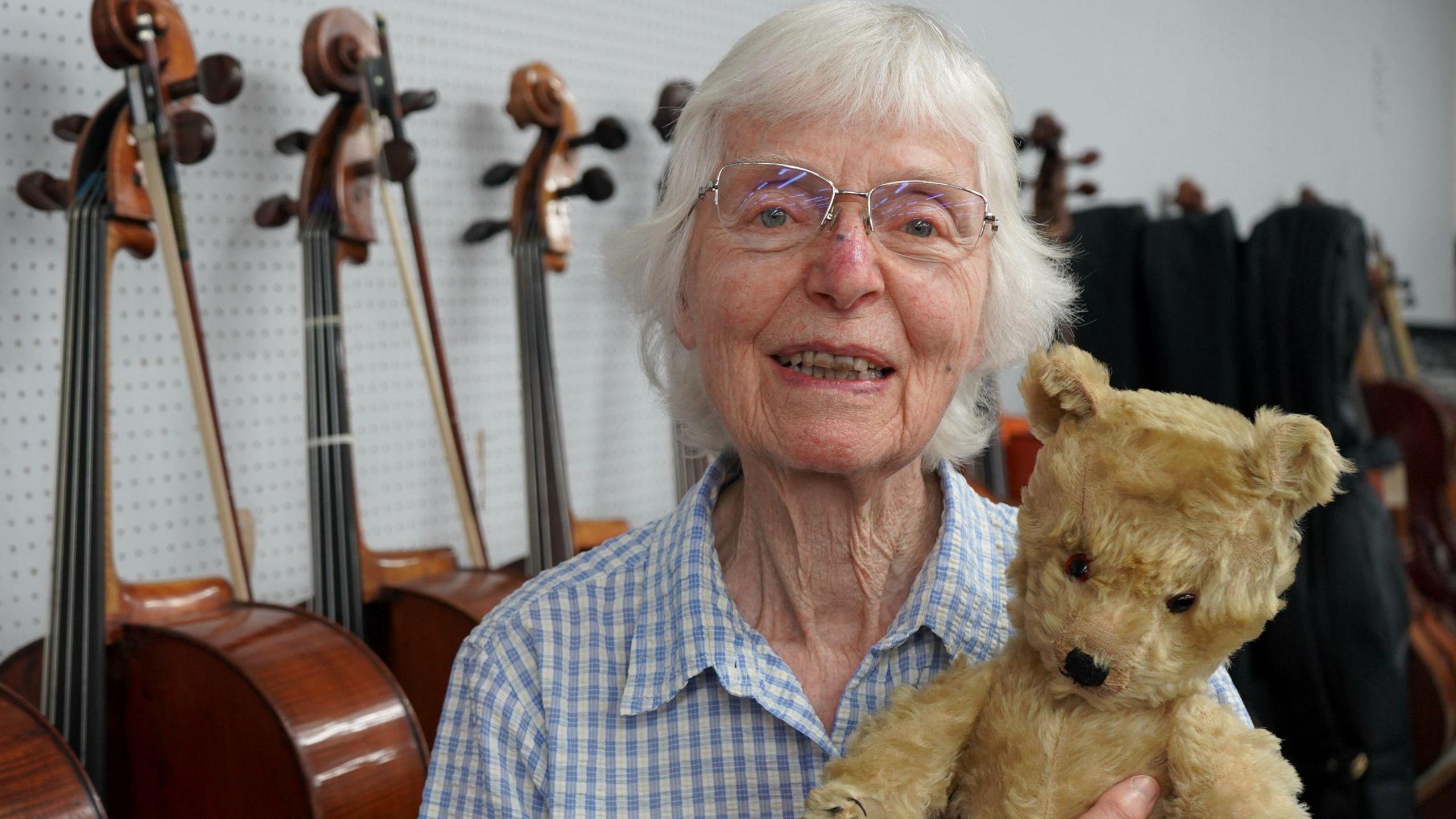 Jaquelin du Pre's sister smiles at the camera and holds an old teddy bear in front of a row of cellos