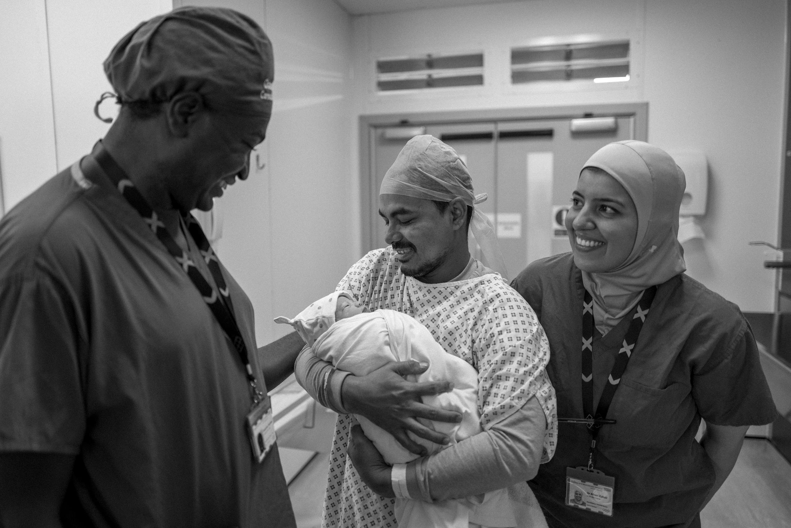 The proud father cradling his newborn, smiling as he shares a moment with two healthcare workers. All are dressed in scrubs