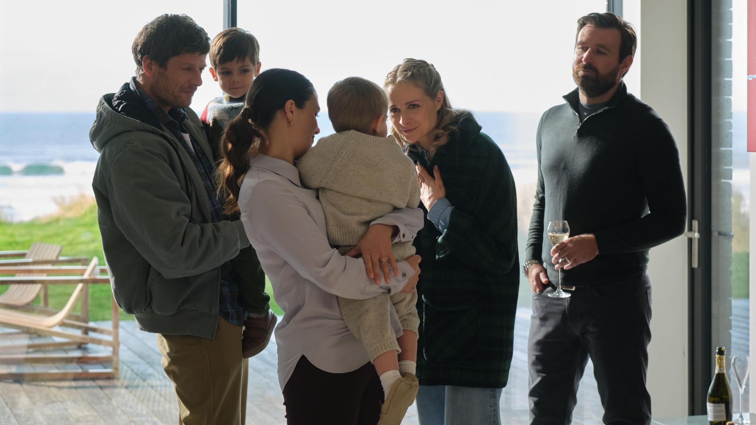 The main characters of Playing Nice are in a room with floor to ceiling windows looking over the beach and sea beyond. The actors are James Norton, Niamh Algar, James McArdle and Jessica Brown Findlay with two young boys.