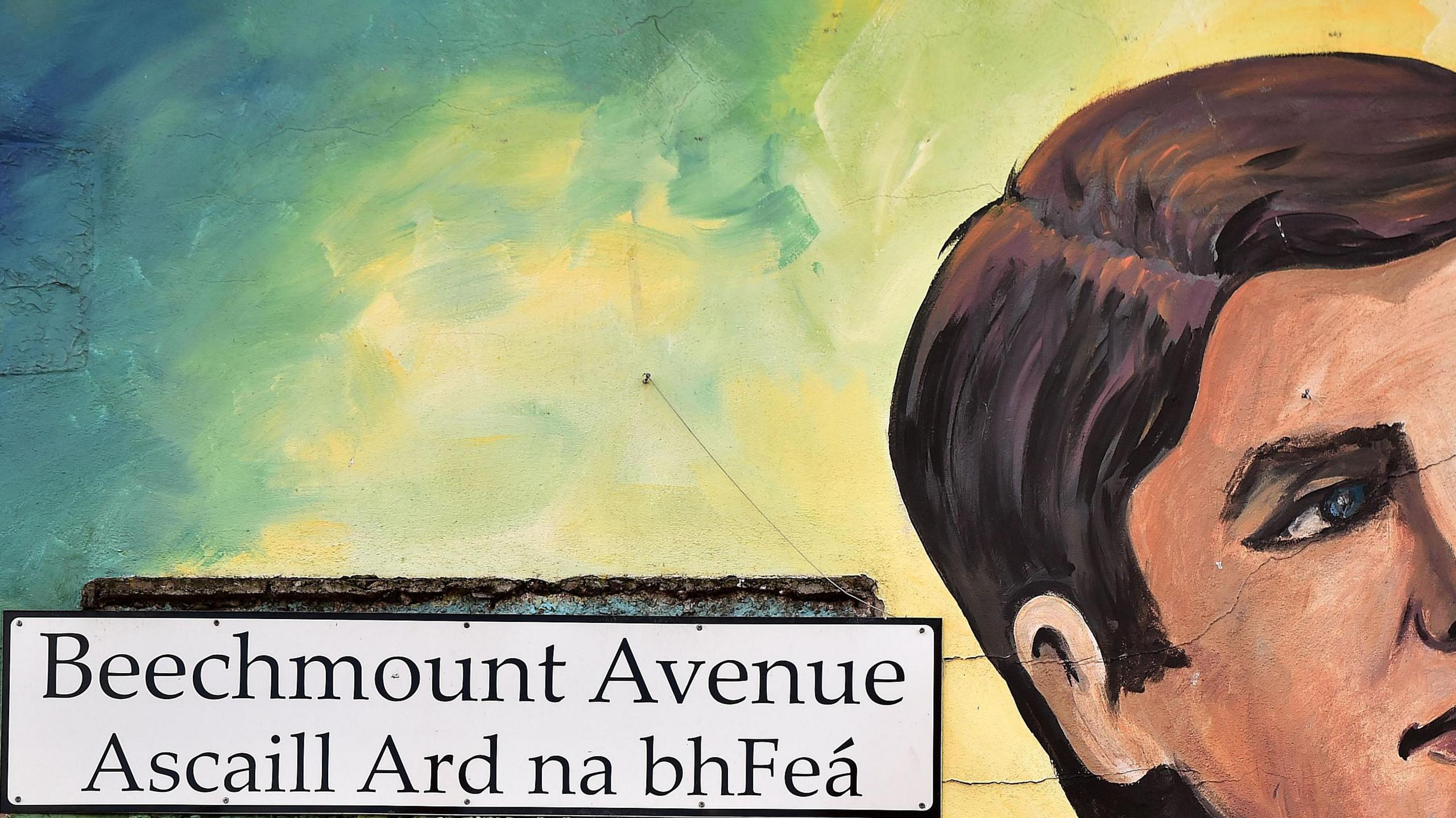 A dual English-Irish street sign for Beechmount Avenue is placed on a wall beside a painted mural of Michael Collins. Under Beechmount Avenue is "Ascaill Ard na bhFeá".