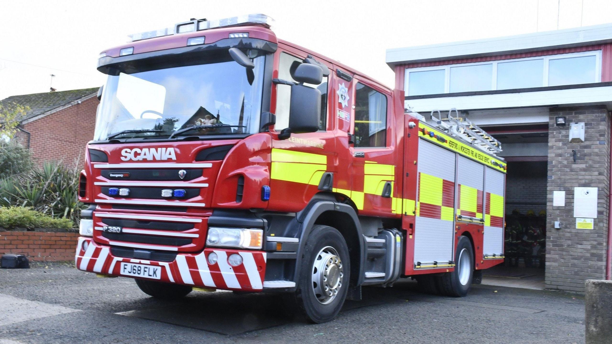 A large fire engine. It is red with bright yellow squares of colour on it, a silver ladder on its roo and blue and white lights on the cab. It is parked in front of an open fire station garage