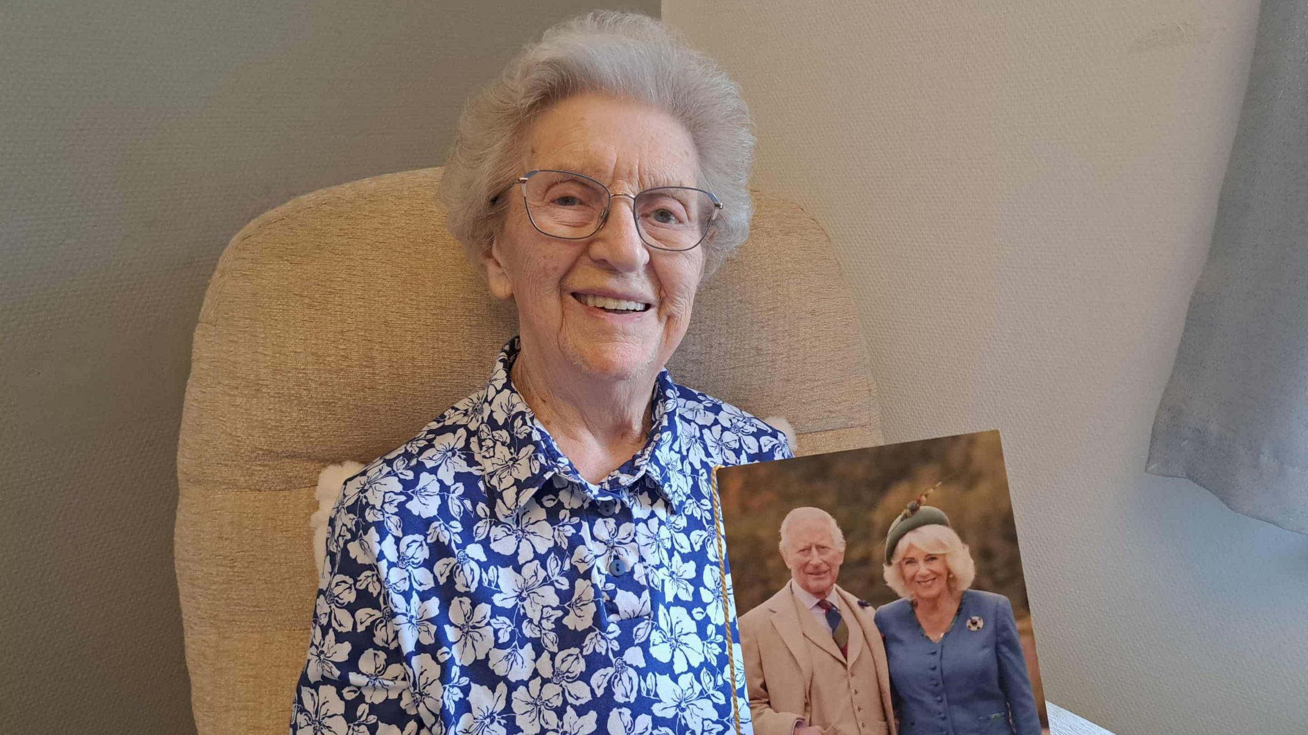 An elderly woman, 100-year-old, Jean Aitken smiles into the camera, holding her 100th birthday card from the King, which has a picture of King Charles and Queen Camilla on it. She has white hair, and is wearing a blue blouse.