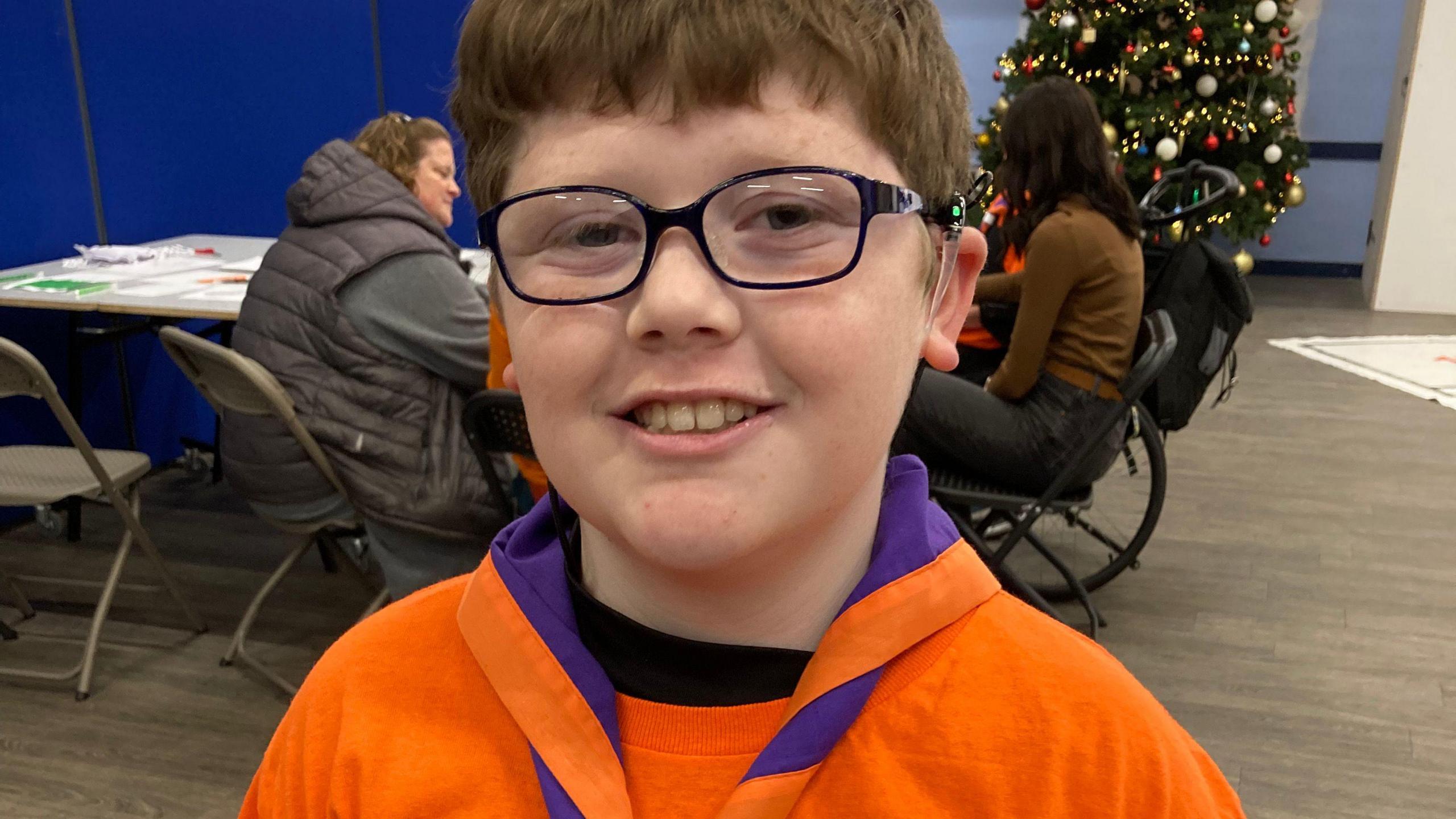 Zack, aged 10, wears a black T-shirt under an orange scouts top. He has short hair and glasses. Behind him, two people are sitting at a table with artwork on it. There is a Christmas tree in the background.