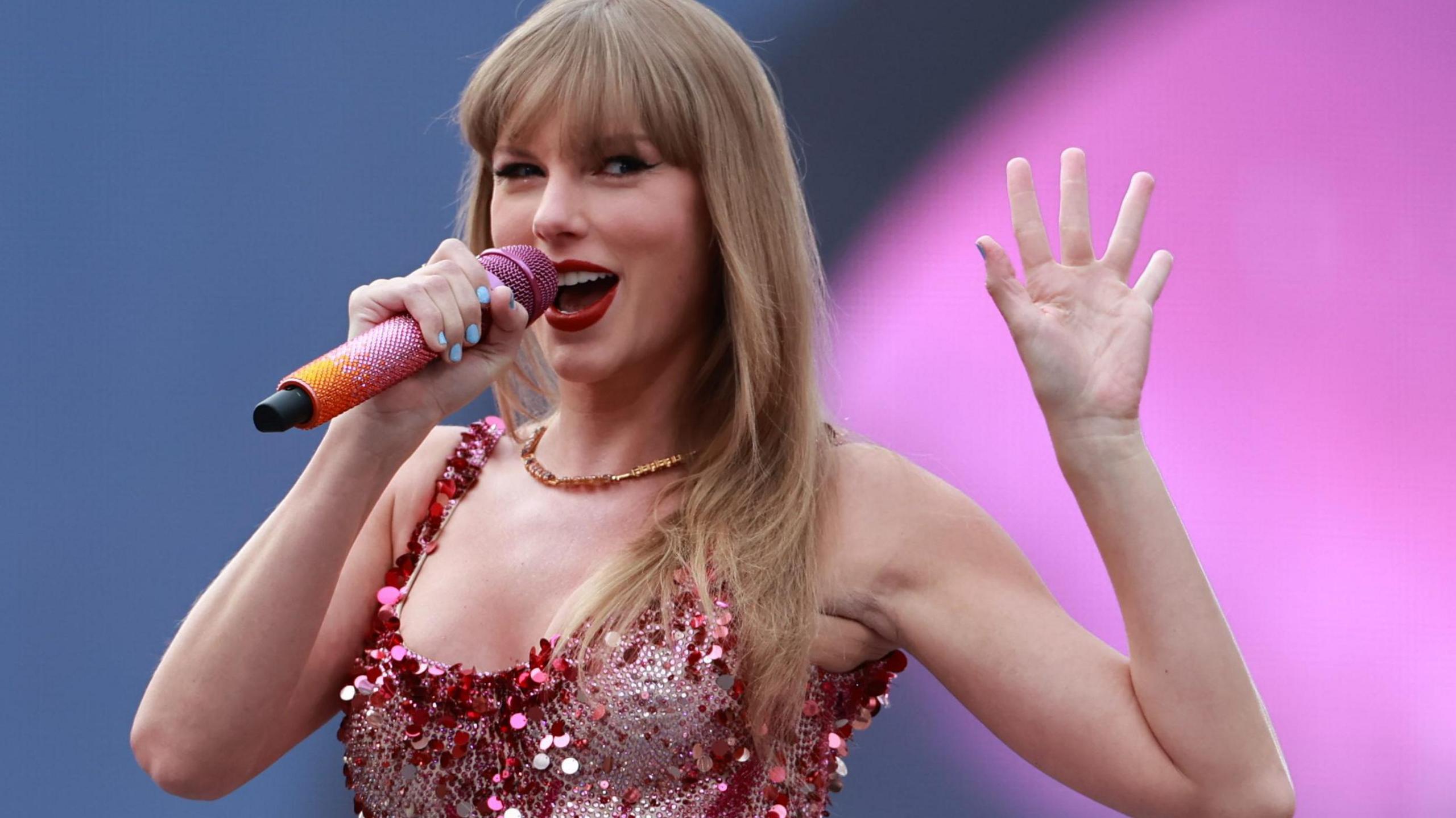 Taylor Swift holds her splayed hand out to the side of her, while clasping an orange and pink diamante microphone in the other. She is wearing a diamante and sequinned pink and silver corset and a gold necklace.