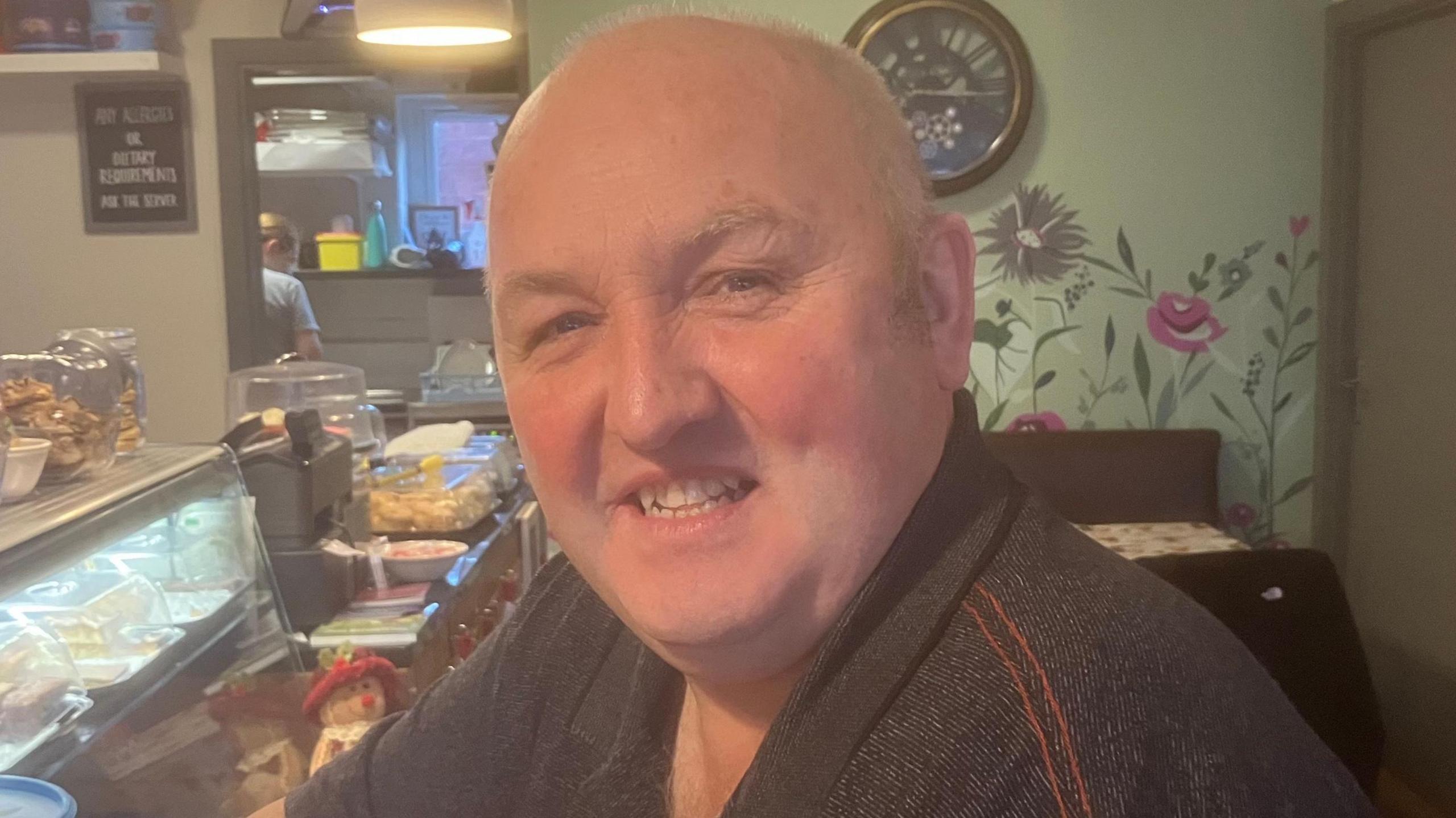 A man wearing a t shirt sits smiling into the camera in a cafe. Behind him is an ornate clock with a table under it. To the side of him are cakes and pastries in a glass refrigerated storage unit.