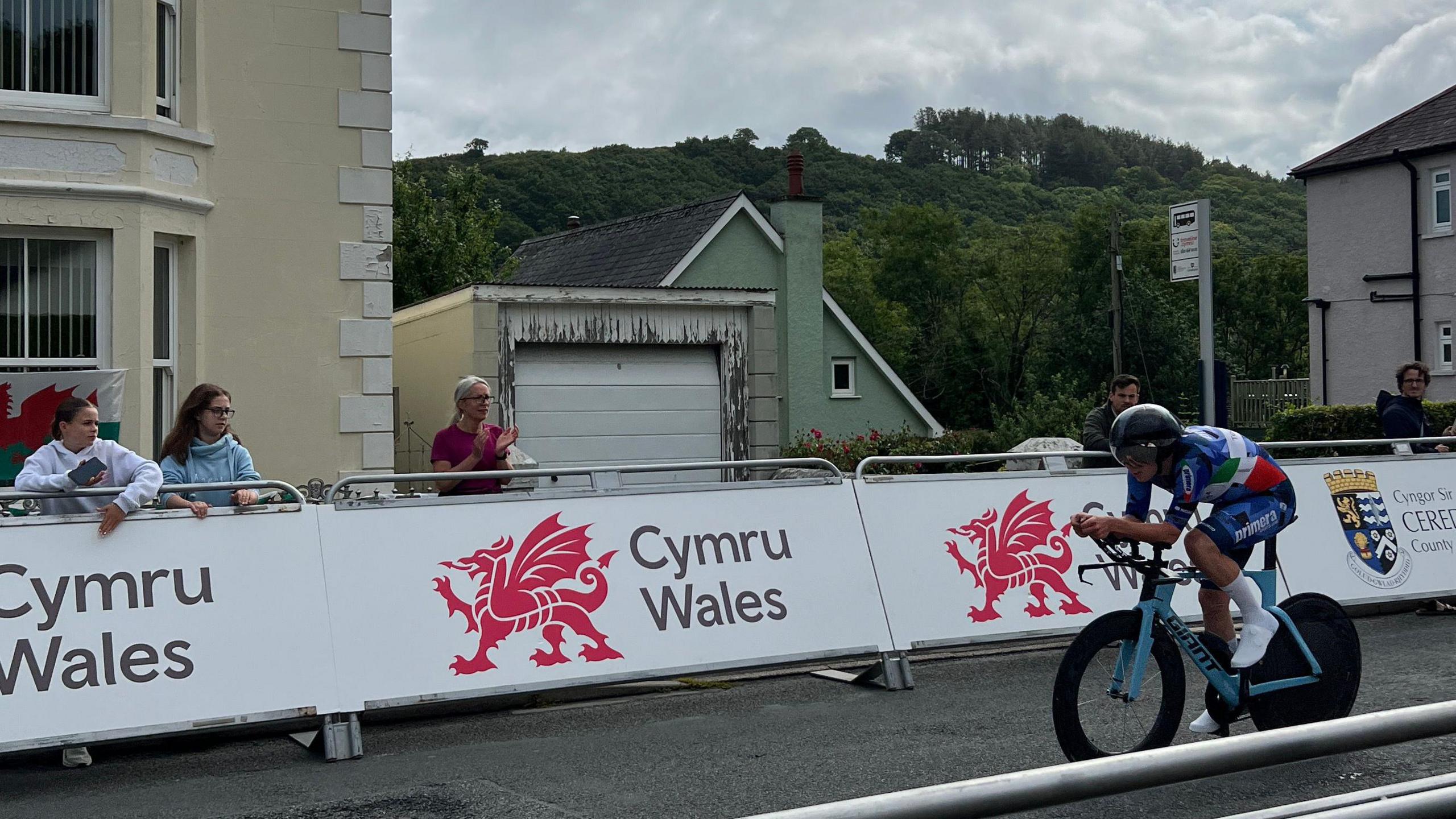 A residential road with barriers which read "Cymru Wales". A few people are stood behind the barrier, watching a cyclist dressed in blue ride by.