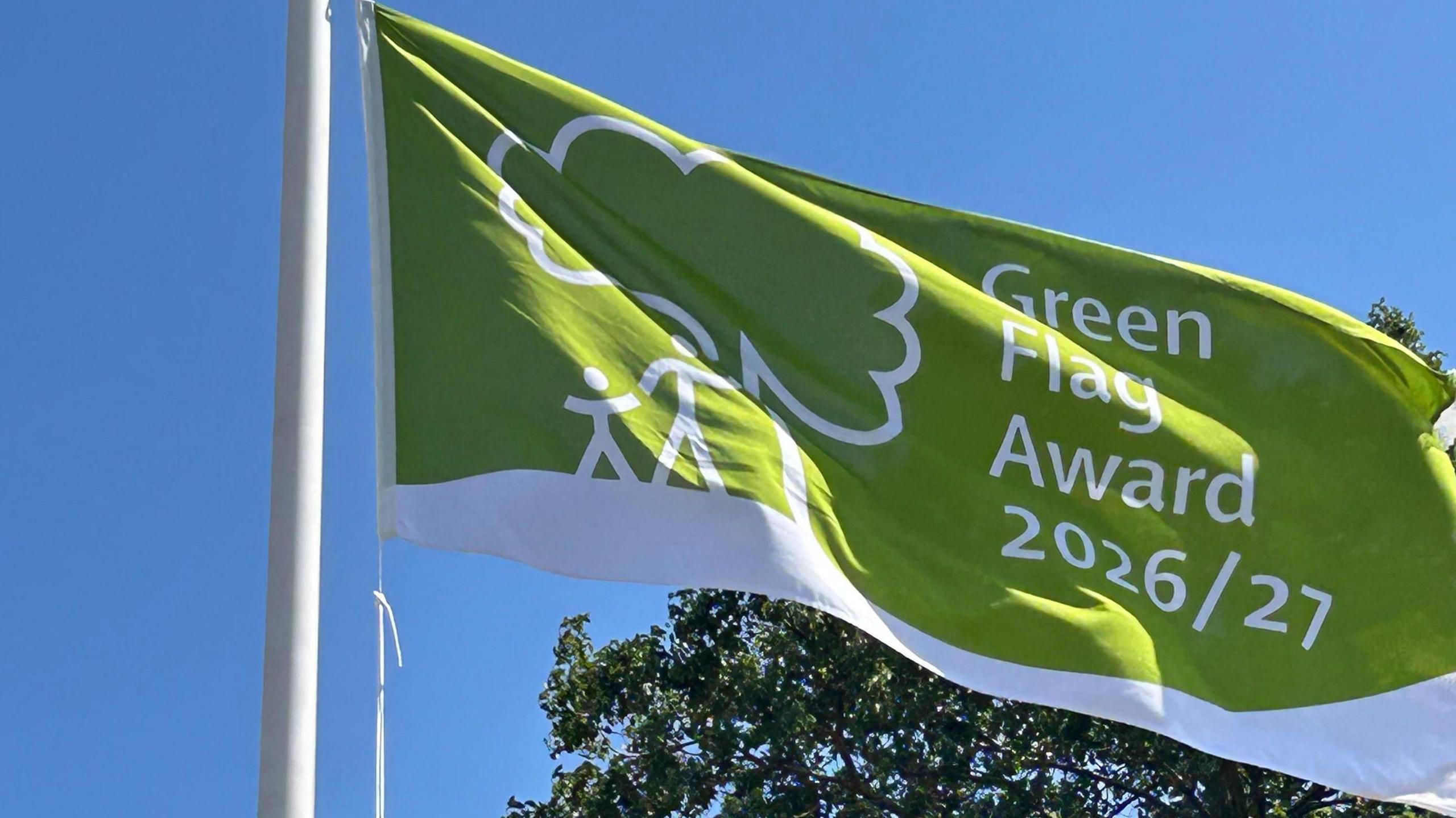 A green flag with a white stripe across the bottom and a tree with a parent and child stood next to it in white. It reads 'Green Flag Award 2026/27. A tree is in the background against the blue sky.