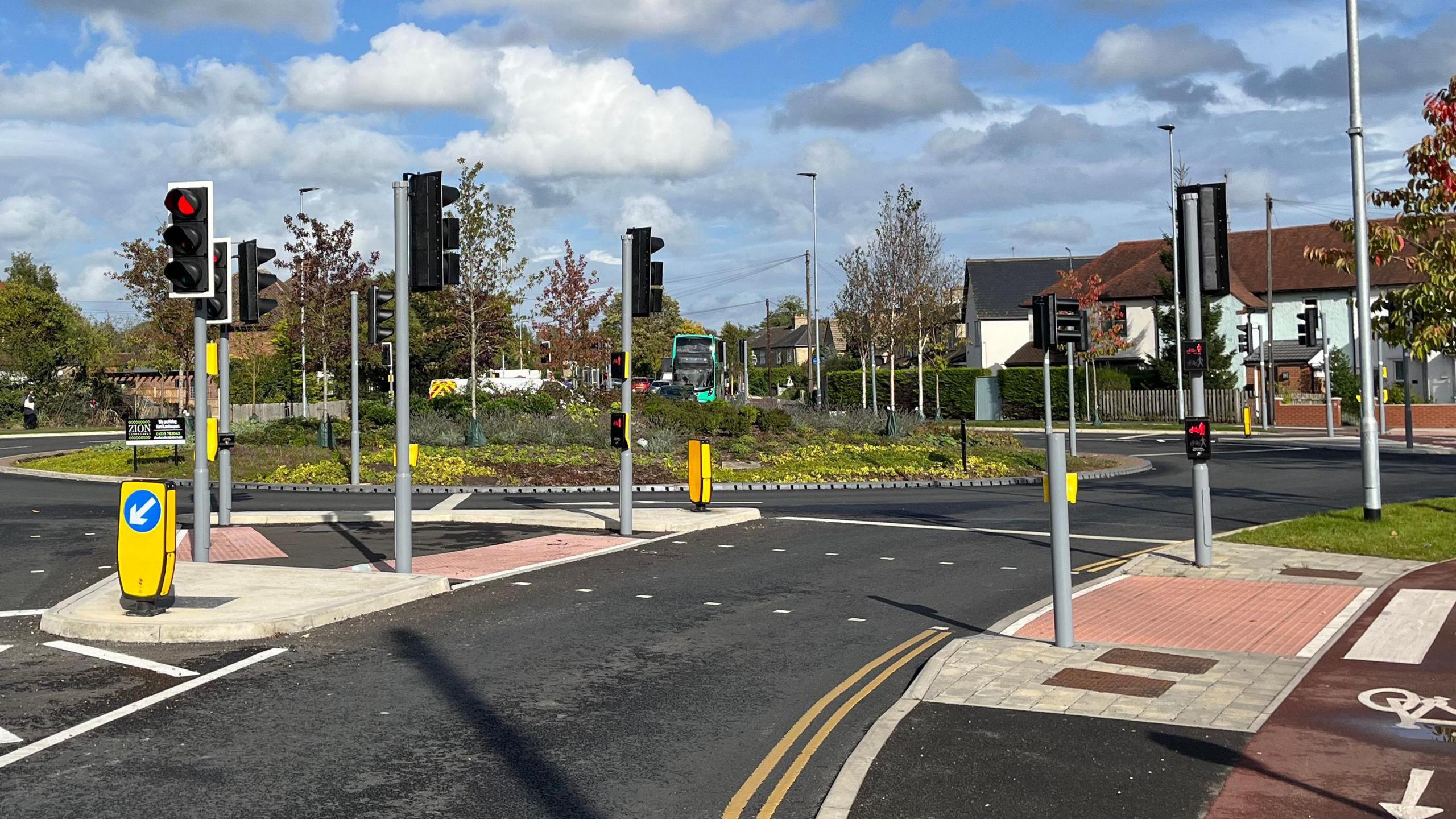 A photograph taken from the road approaching a roundabout. A number of sets of traffic lights can be seen, and houses and a green bus can be seen in the distance. 