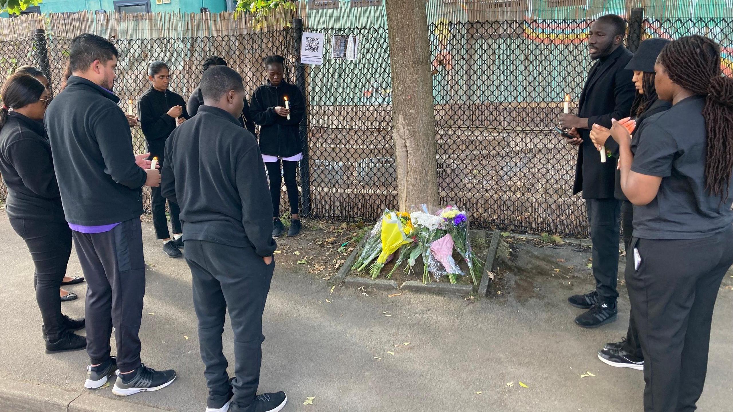 A group of people in mourning gather around a tree, at the bottom of which they have placed bouquets of flowers.