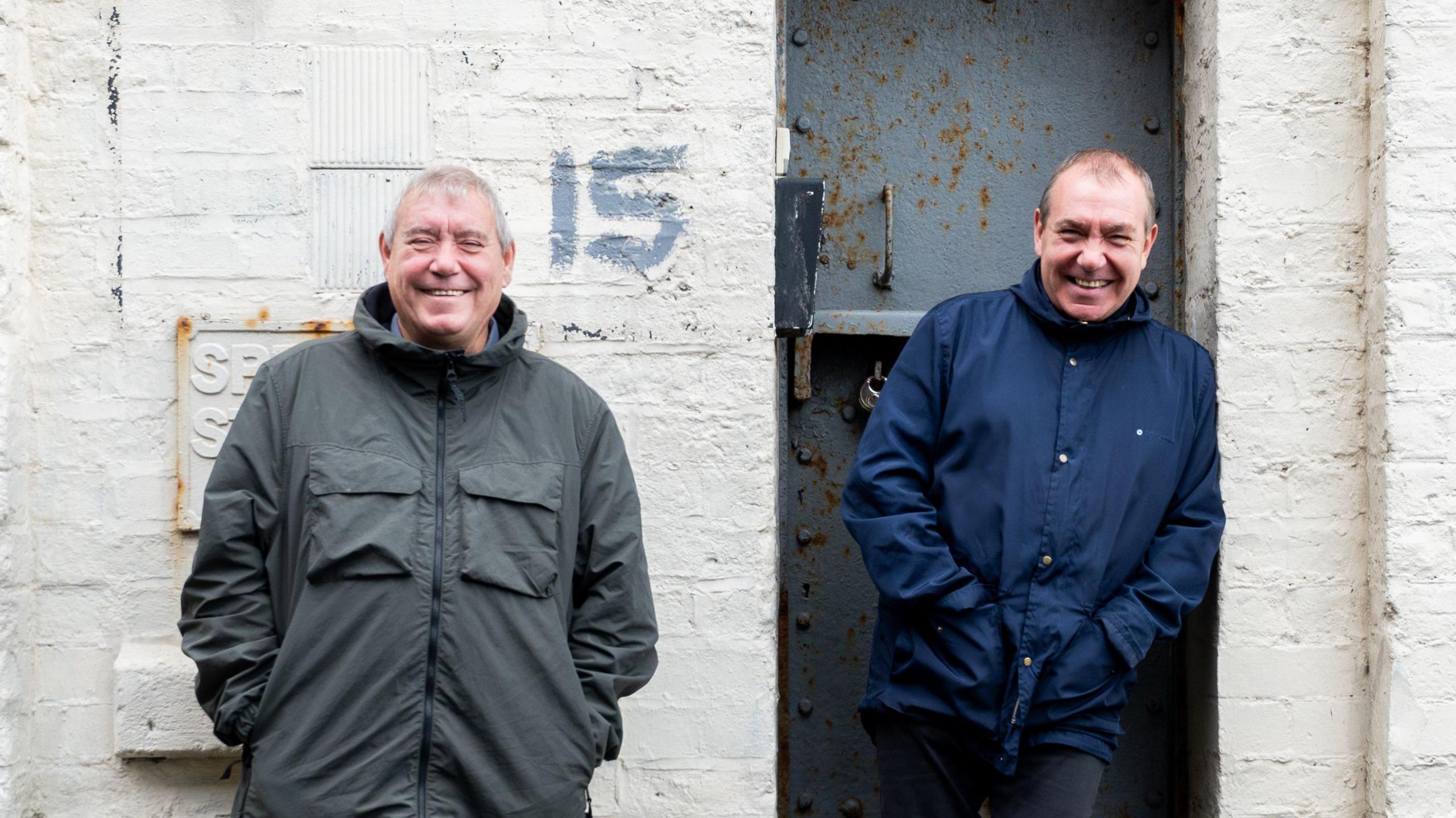 Tony and Chris Griffiths outside their studio in Birkenhead. Tony leans against a white brick wall while Chris sits on the door step. 