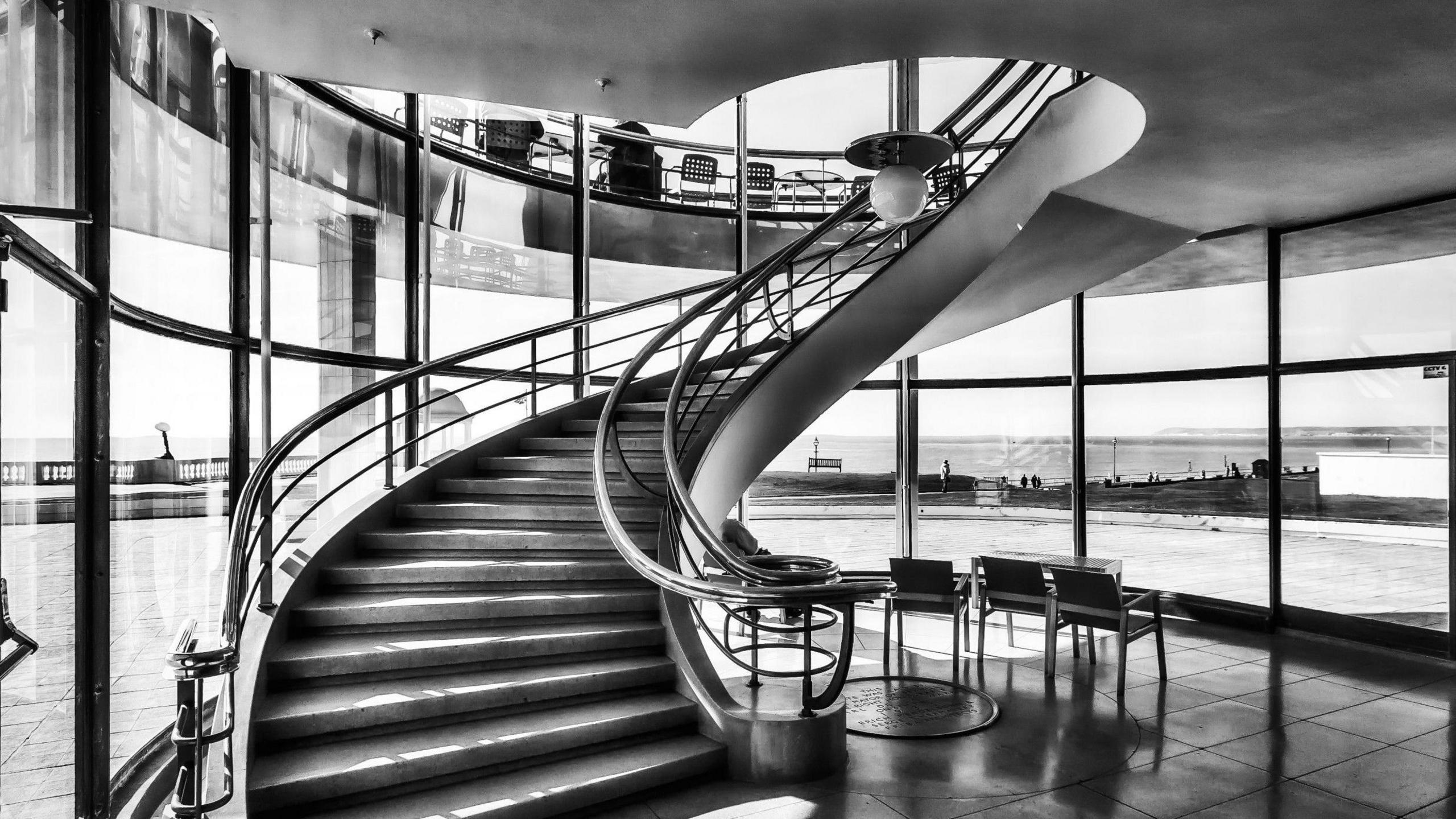 A black and white photograph of a spiral staircase inside the De La Warr Pavilion, with a set of steel chairs at the base, looking out to sea through floor-to-ceiling windows.