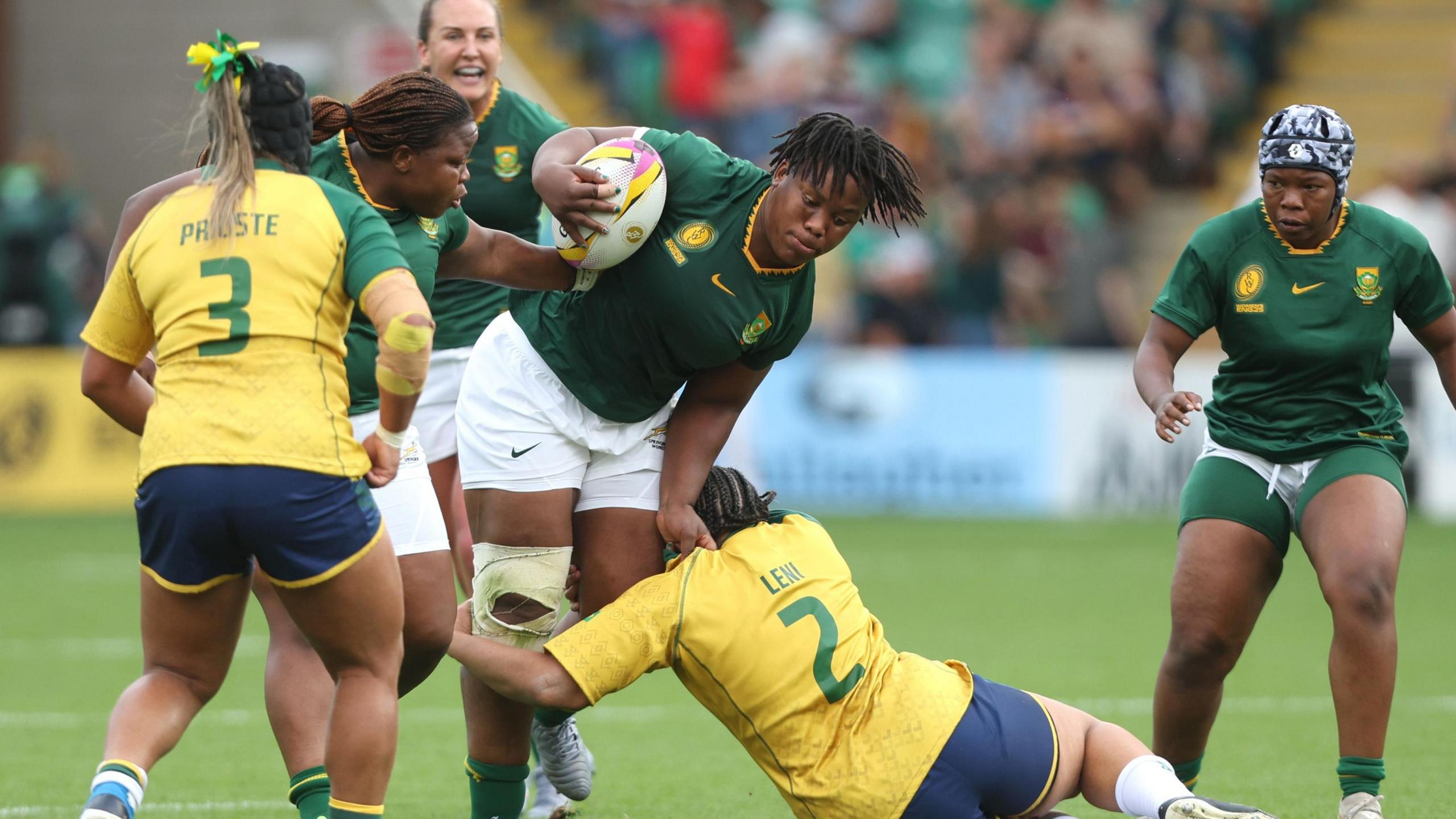Babalwa Latsha, wearing a green South Africa rugby shirt, white shorts and a white brace on her right knee, holds a rugby ball in the crook of her right arm as she goes through an attempted knee-high tackle by a Brazilian opponent lying on the floor, who is wearing a yellow shirt with the number two on its back. One other Brazilian, with the number three shirt, looks on as three South Africa team-mates are seen around Latsha during a pool match at the 2025 Women's Rugby World Cup