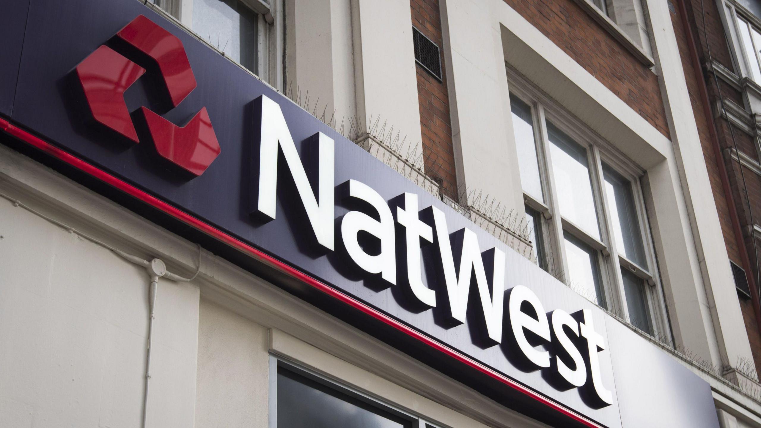 Signage above a NatWest bank branch. The logo is a set of three red chevrons arranged in a triangular shape, and the lettering is in white. Both are on a purple backboard.